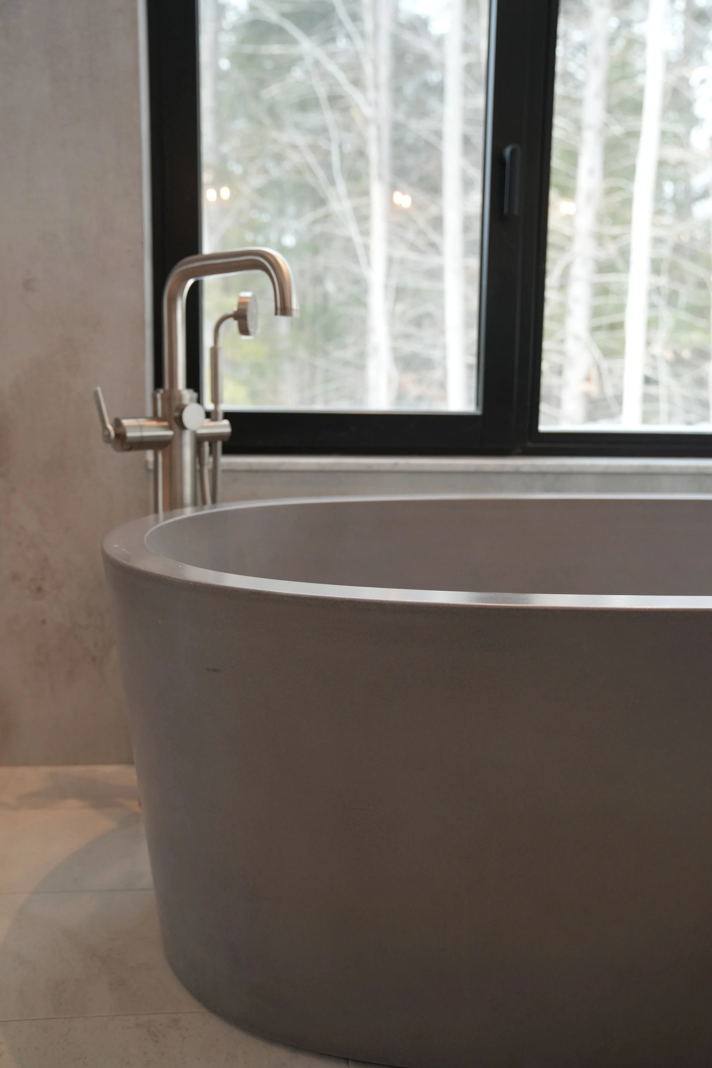 A modern white bathtub with a curved edge in front of a large window showing a view of trees outside. A silver faucet is mounted on the side of the tub.
