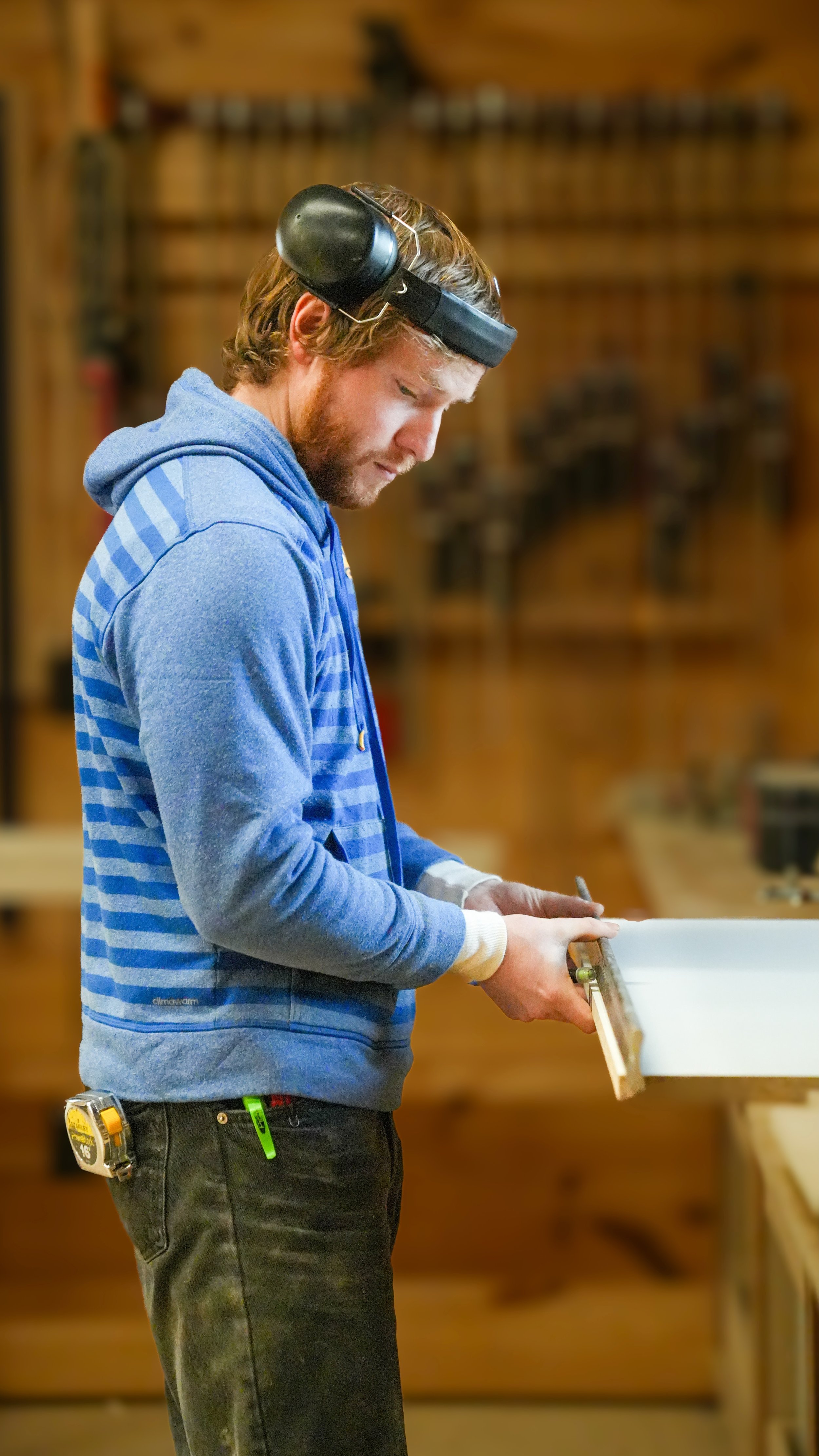 A man with a beard wearing a blue hoodie and black pants is holding a woodworking tool in a workshop. He wears safety earmuffs on his head and appears focused on his work.