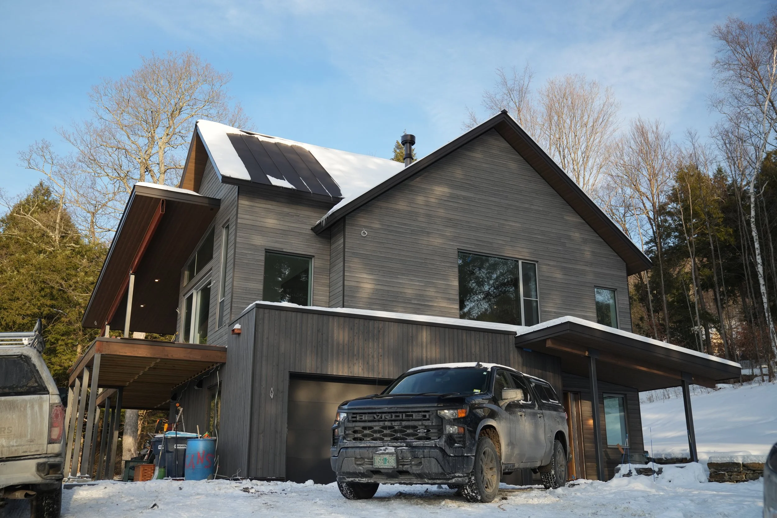 A modern gray house with large windows and a sloped roof partially covered with snow, surrounded by trees and parked vehicles on a snowy ground under a partly cloudy sky.