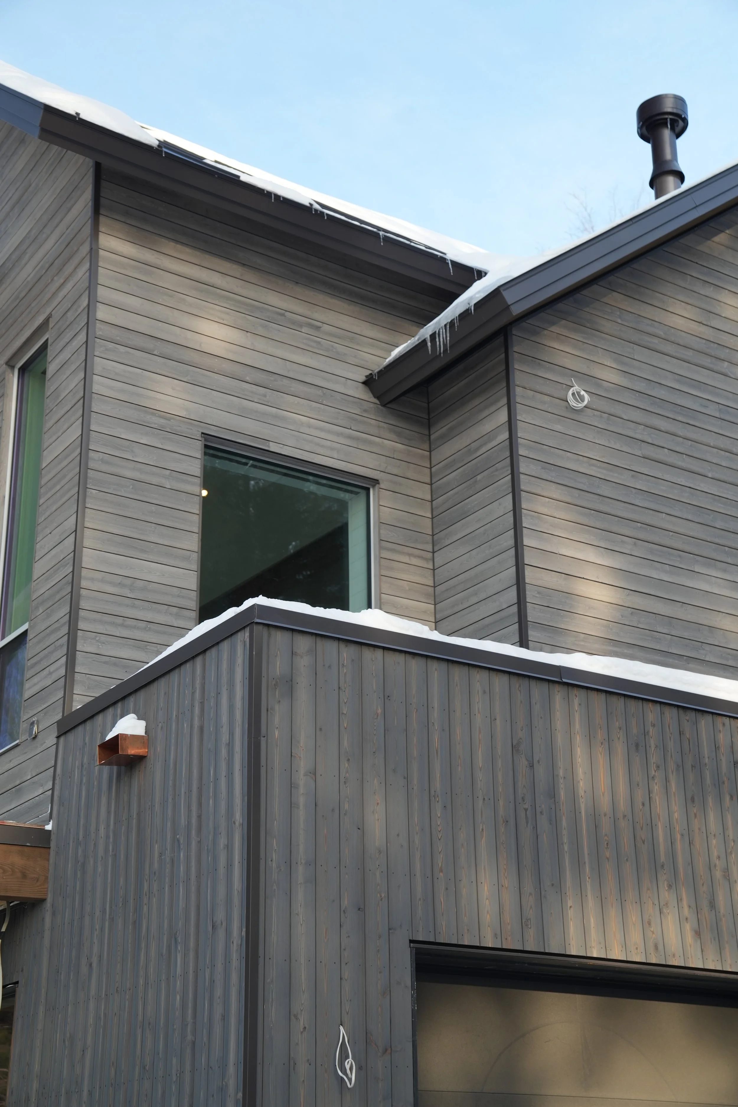 A modern house with wood siding, snow on the roof, icicles hanging from the eaves, and a chimney emitting no smoke.