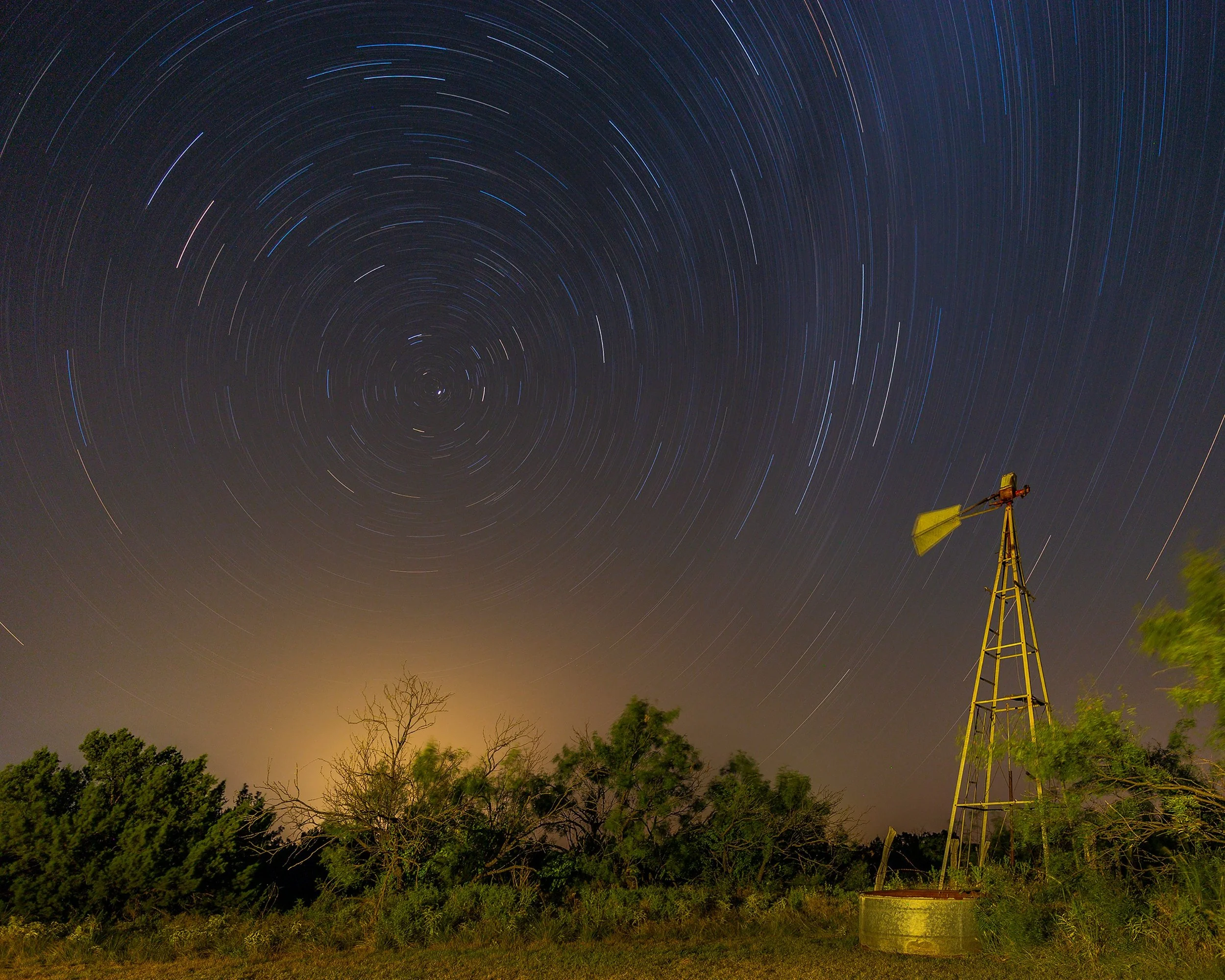 Night sky with star trails circling around the celestial pole, trees and a windmill in the foreground.