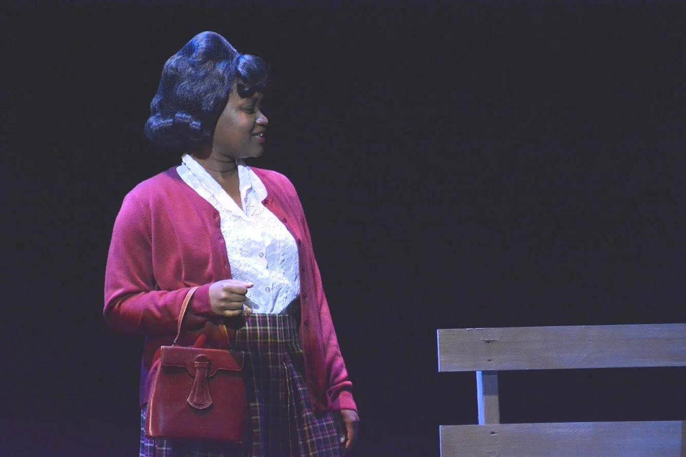 A woman standing on stage in front of a dark background, wearing a red cardigan, white blouse, plaid skirt, and carrying a red purse.