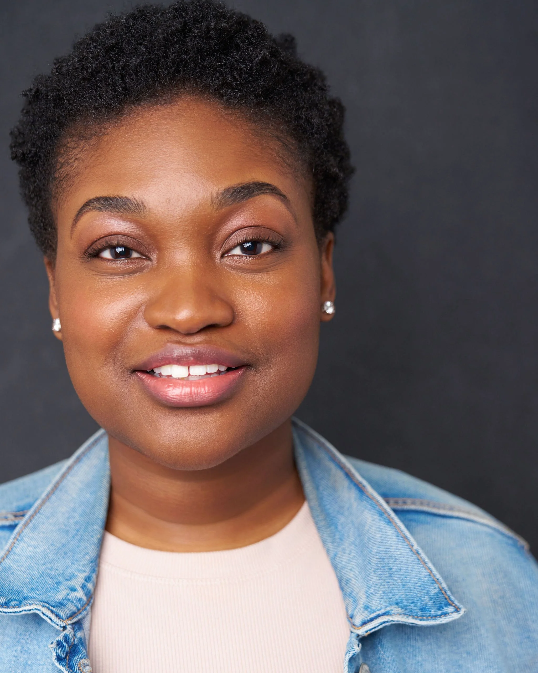 Close-up of a woman with short, curly black hair, wearing earrings, a white top, and a denim jacket, smiling against a dark background.