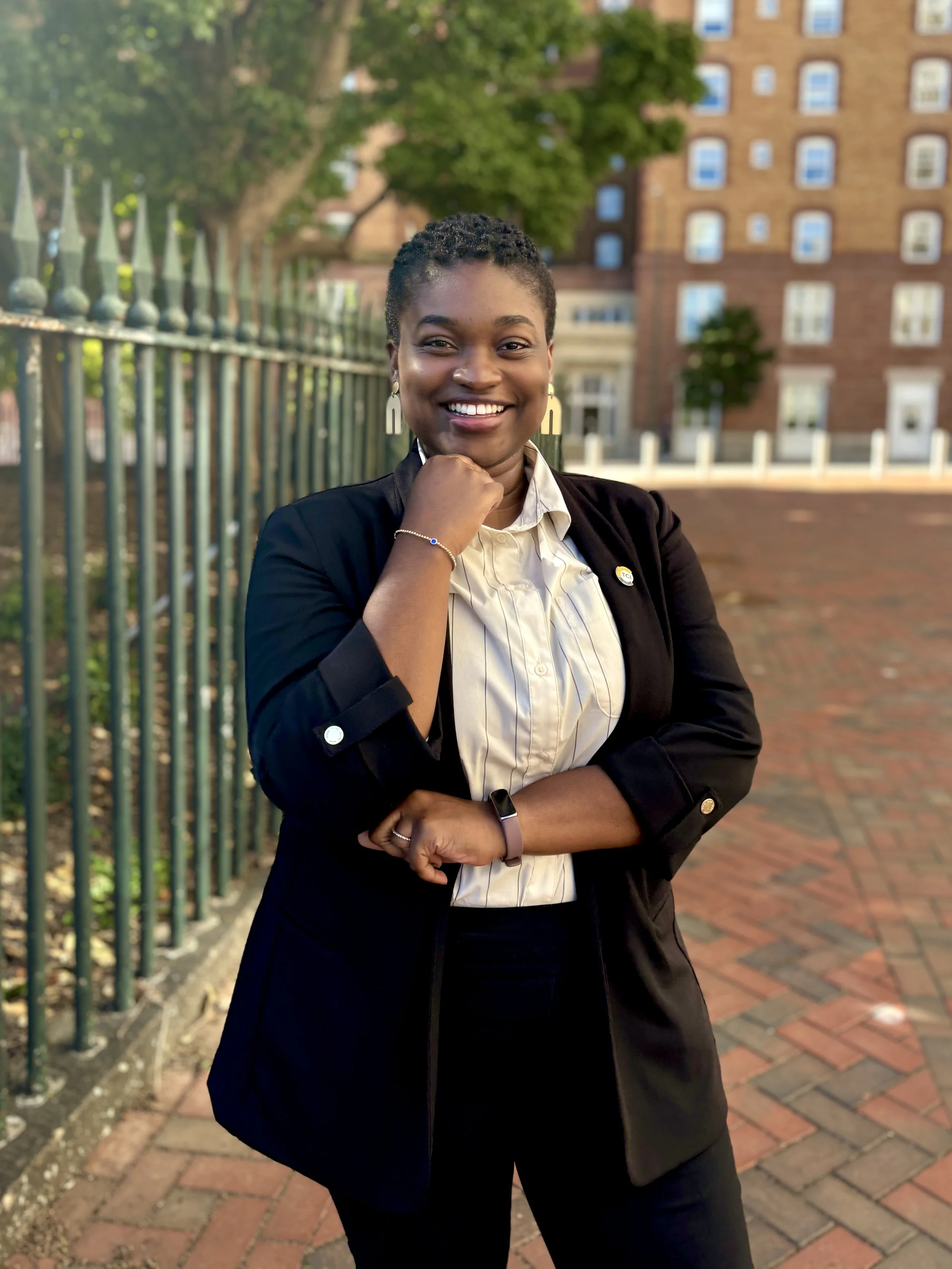 A confident African American woman in formal attire, standing outdoors near a decorative metal fence with a brick building and trees in the background.