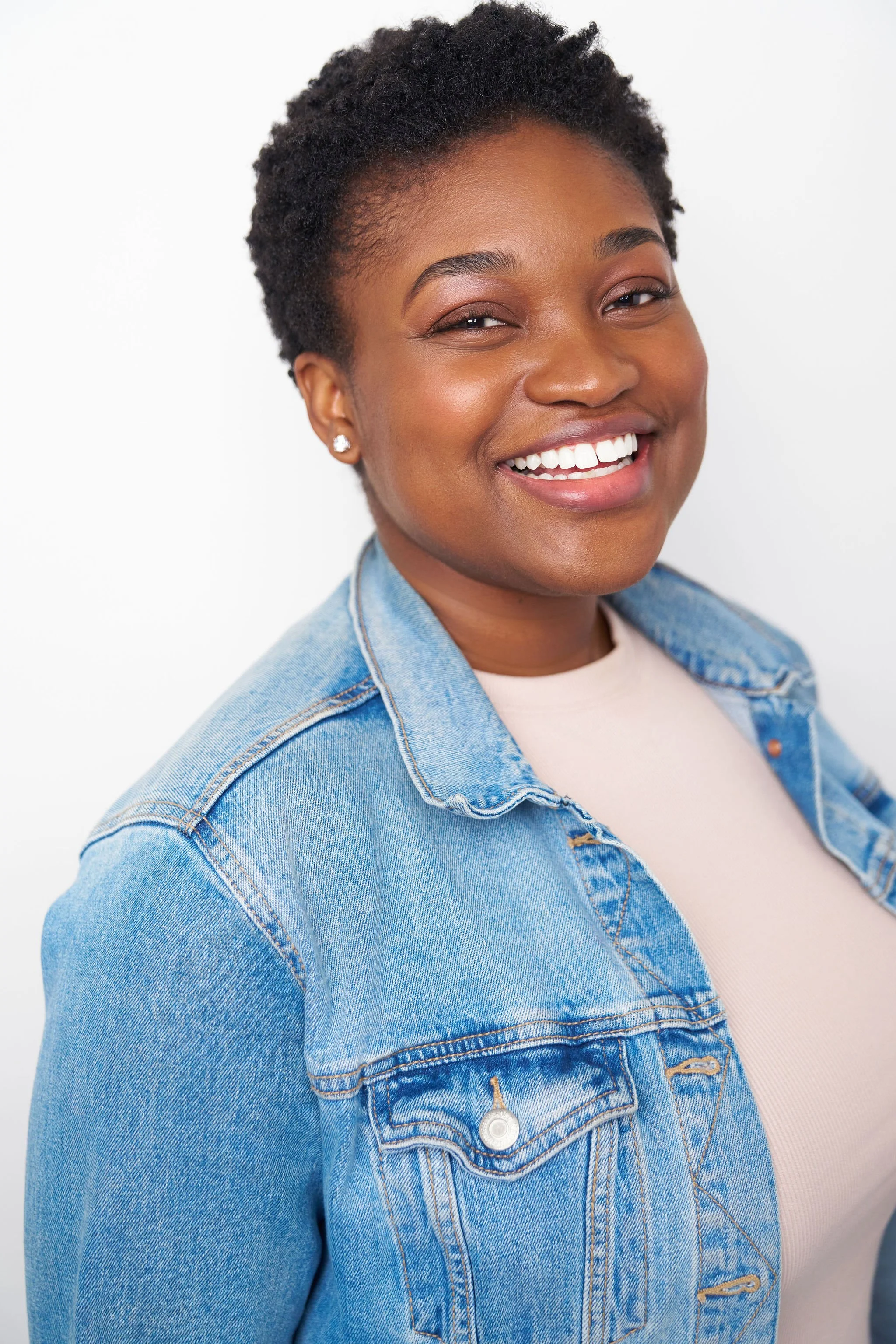 Close-up of a smiling Black woman with short curly hair, wearing a denim jacket and a white shirt, standing against a white background.
