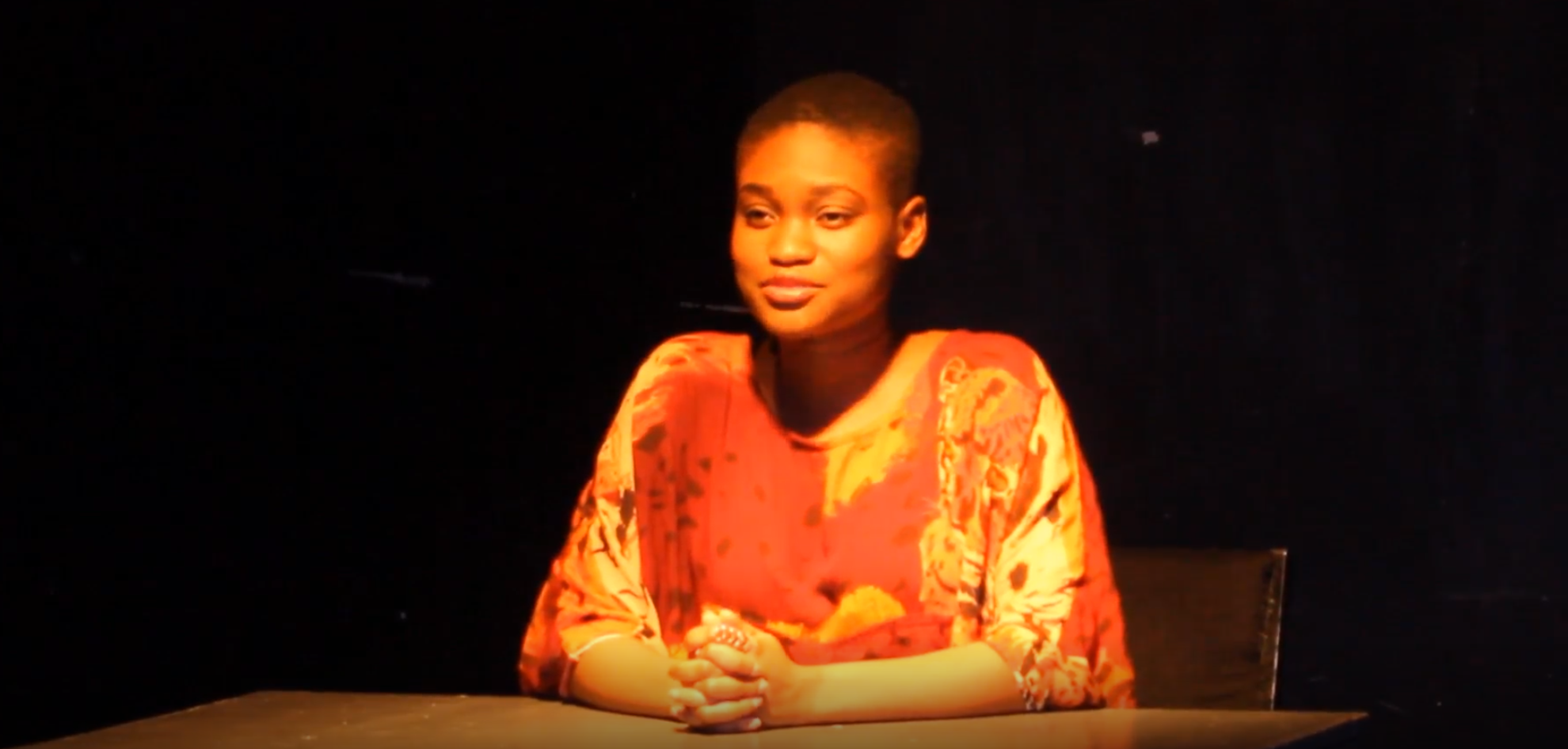 A woman with short hair and a patterned orange and red top sitting at a table against a dark background.