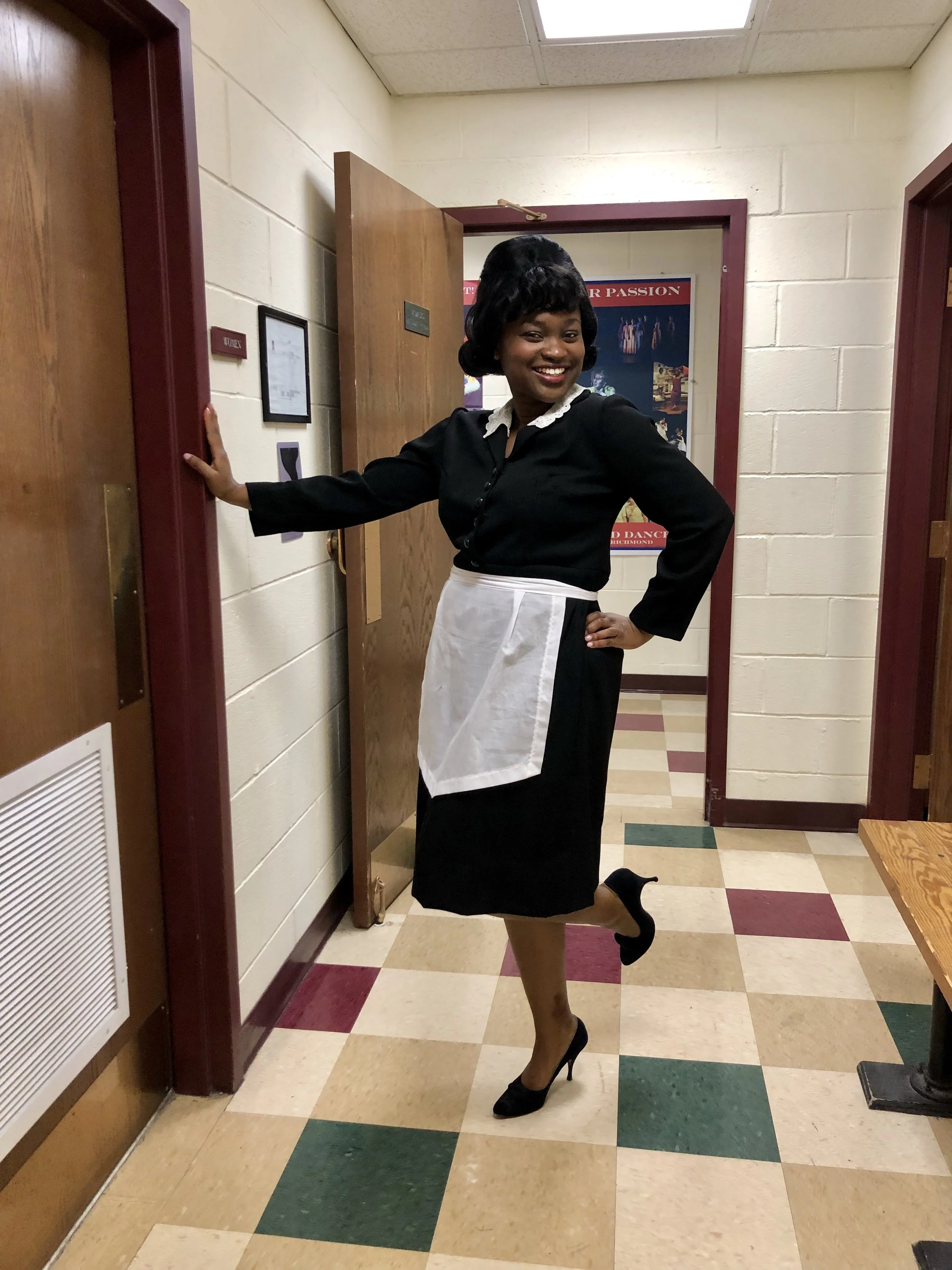 A woman in a black dress with white collar and apron, black high heels, and vintage hairstyle, is posing with one hand on a wall, smiling, in a hallway with multicolored tiled floor and open doors.