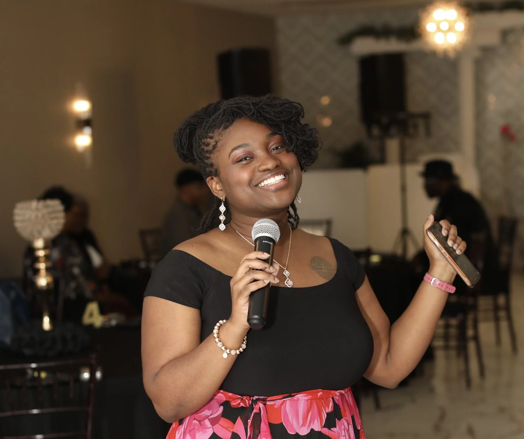 A smiling woman with styled dreadlocks holding a microphone and a mobile phone at an indoor event, wearing a black top, pink floral skirt, jewelry, and a pink wristband.