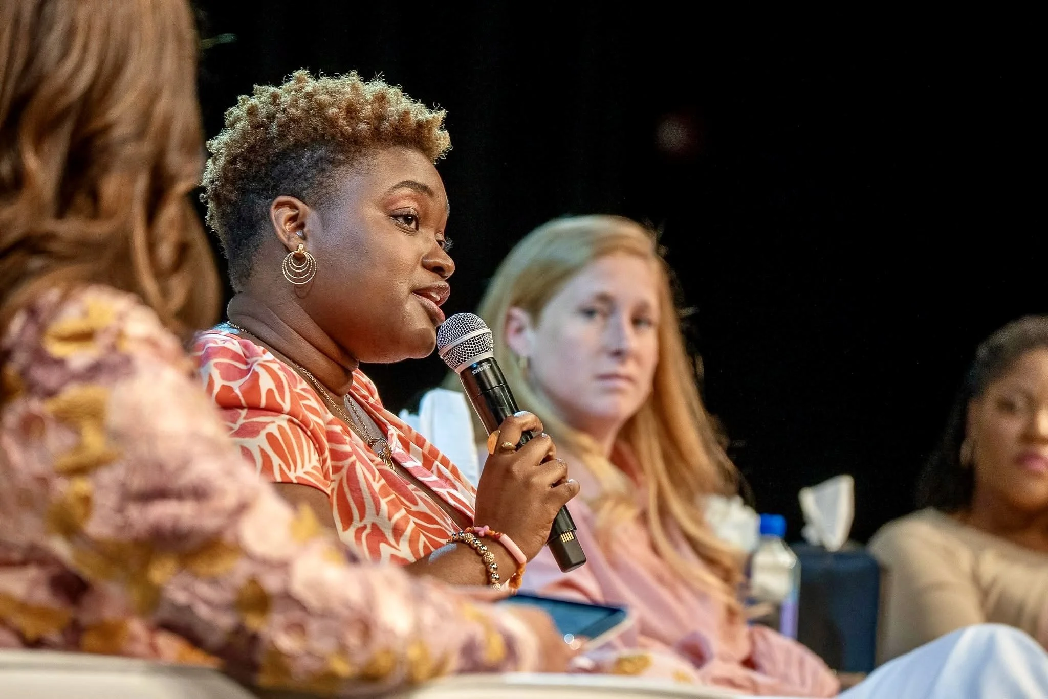 A woman with short curly hair and hoop earrings speaking into a microphone during a panel discussion, seated next to other women.