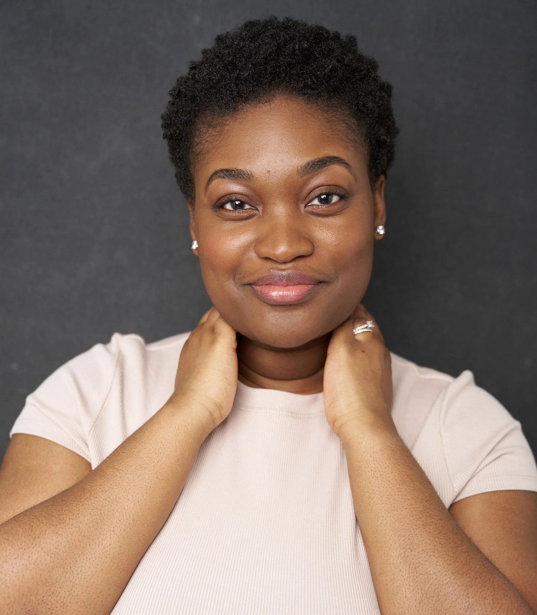Close-up of a smiling African American woman with short curly hair, wearing pearl earrings and a light-colored shirt, against a dark background.