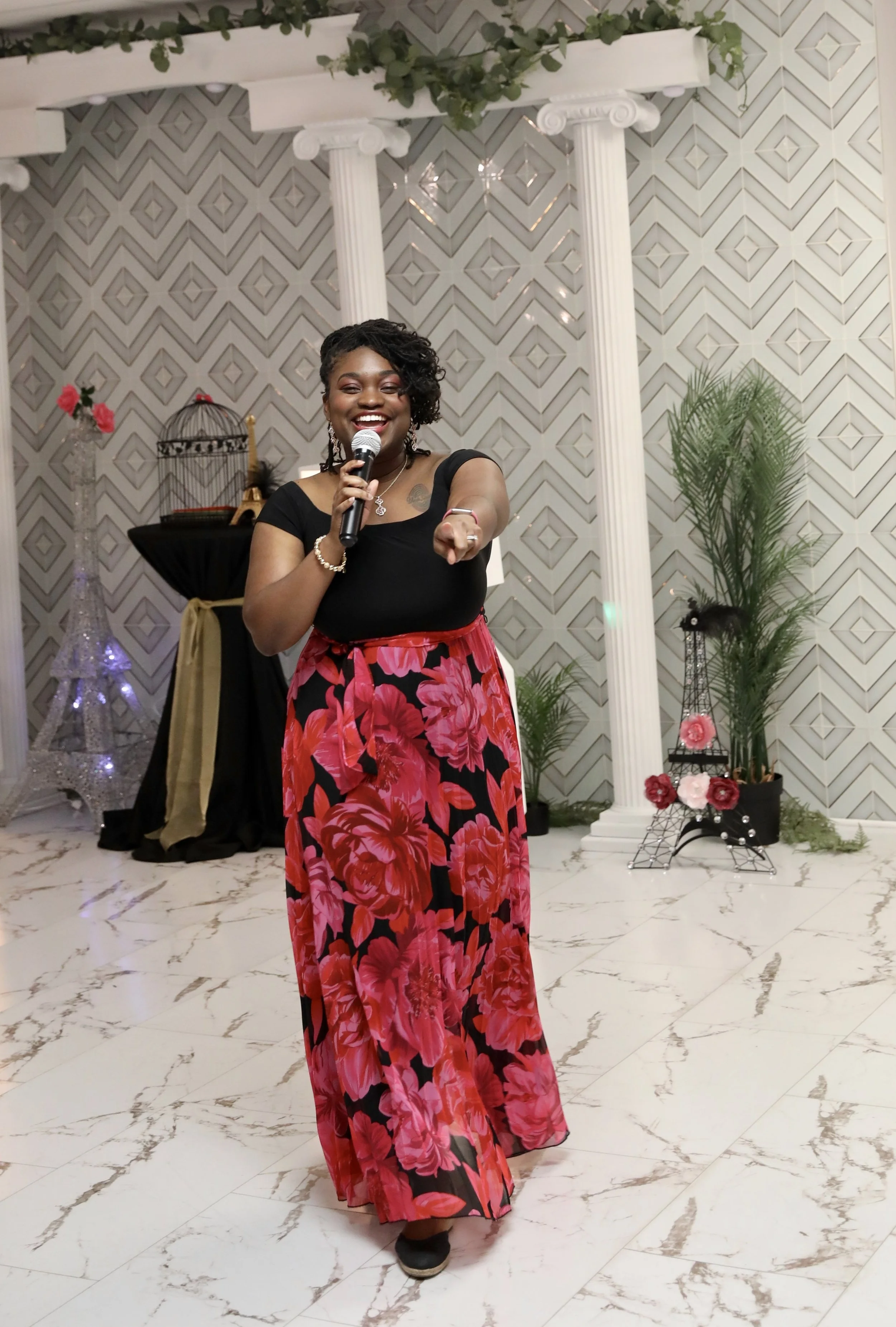 A woman with dark skin, black curly hair, and a broad smile is holding a microphone, pointing forward, and wearing a black top and a long red floral skirt. The background features classical white columns, green plants, and decorative elements like a birdcage, a small Eiffel Tower, and pink flowers in pots, set on marble flooring.