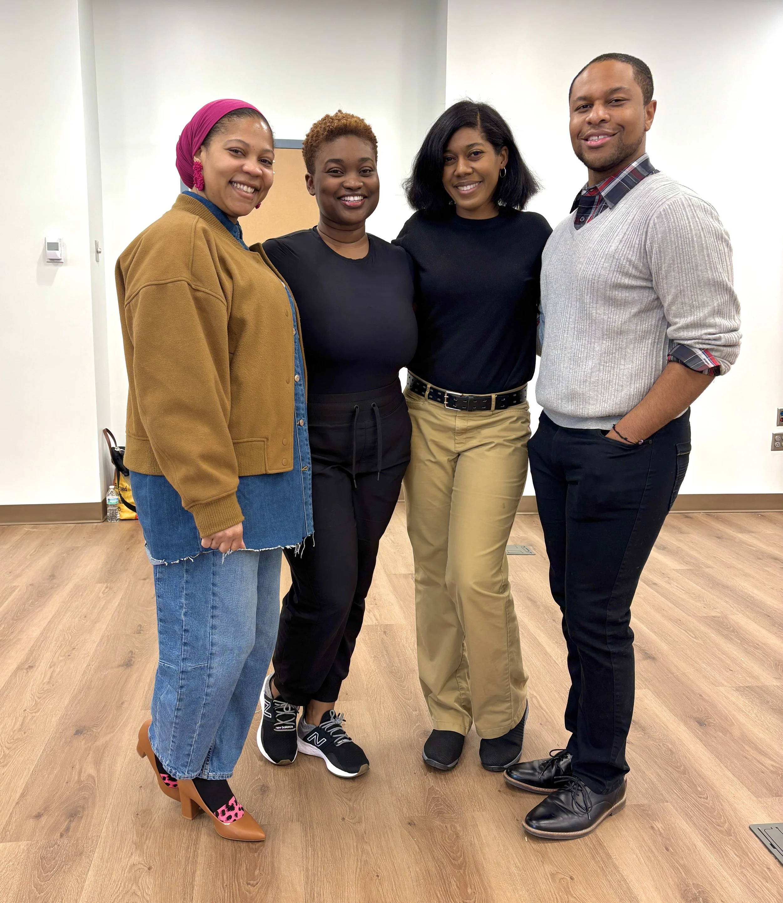 Group of four diverse adults smiling in an indoor room with wooden floor and plain white wall, dressed in casual clothing.