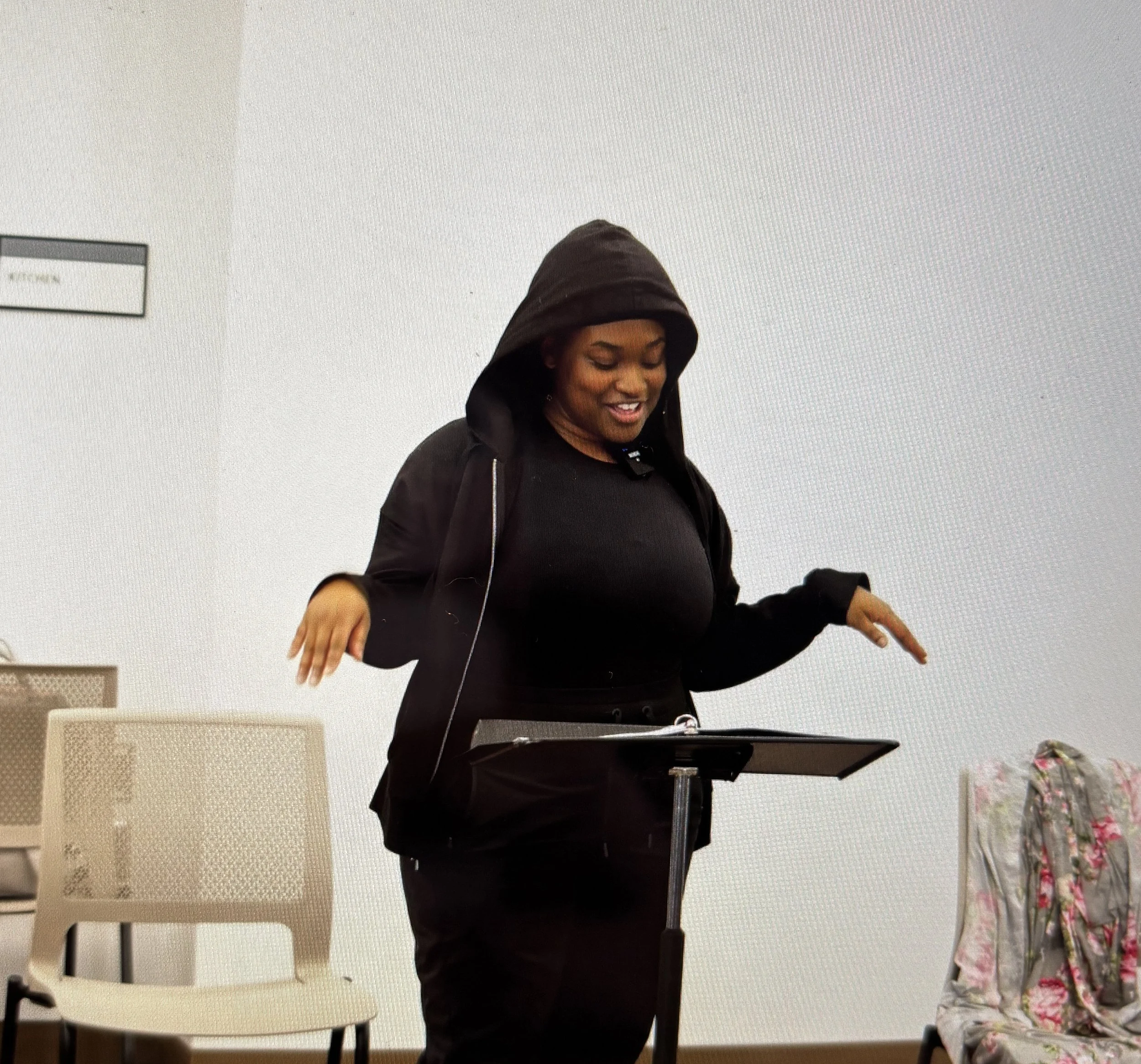 A woman wearing a black hoodie and black jacket, smiling while looking down, standing in front of a music stand in a cozy room with chairs, a floral tablecloth, and a floral pillow on a chair.
