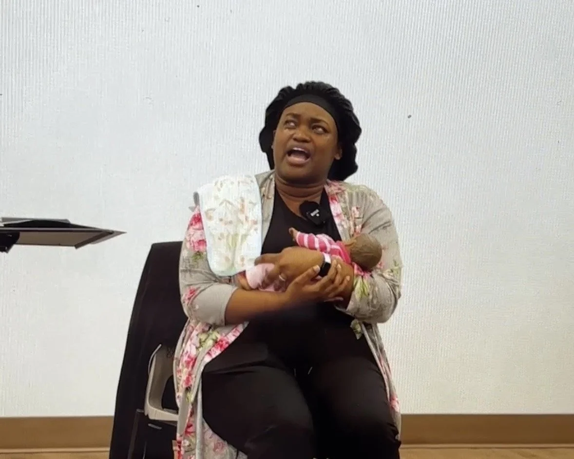 A woman sitting in a chair holding a baby, possibly breastfeeding, with a look of surprise or discomfort on her face.