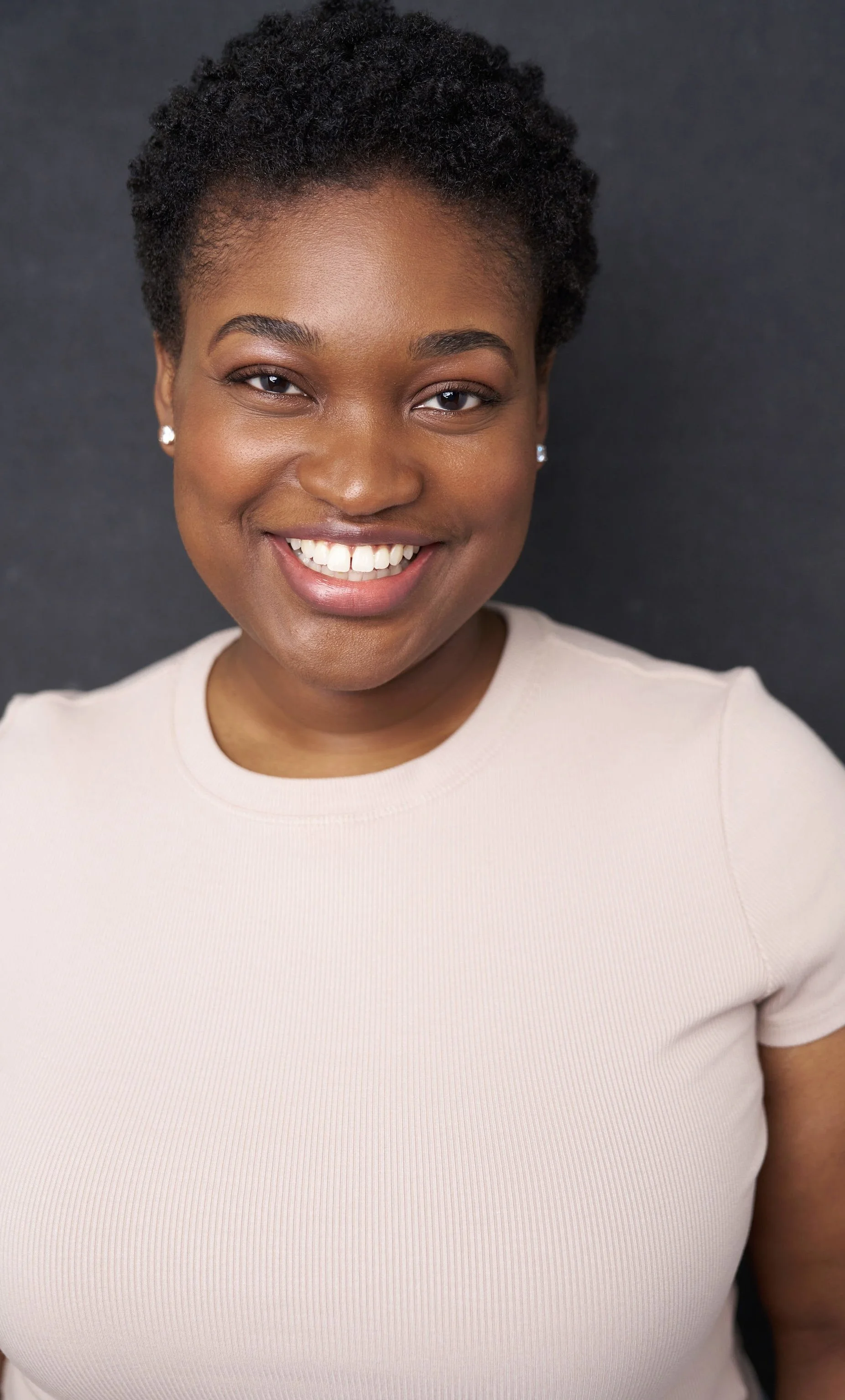 Close-up portrait of a smiling black woman with short natural hair, wearing pearl earrings and a beige top against a dark background.