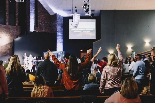 People gathered in church or auditorium, some with hands raised during a worship service or event.