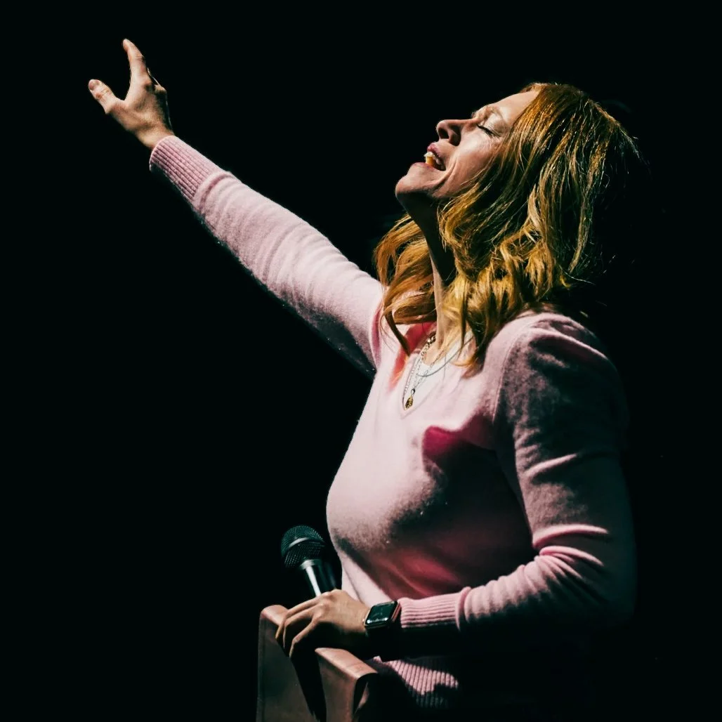 A woman with reddish-brown hair, wearing a pink sweater, holding a microphone, with her eyes closed and arm raised, appearing to be singing or speaking passionately in a dark setting.