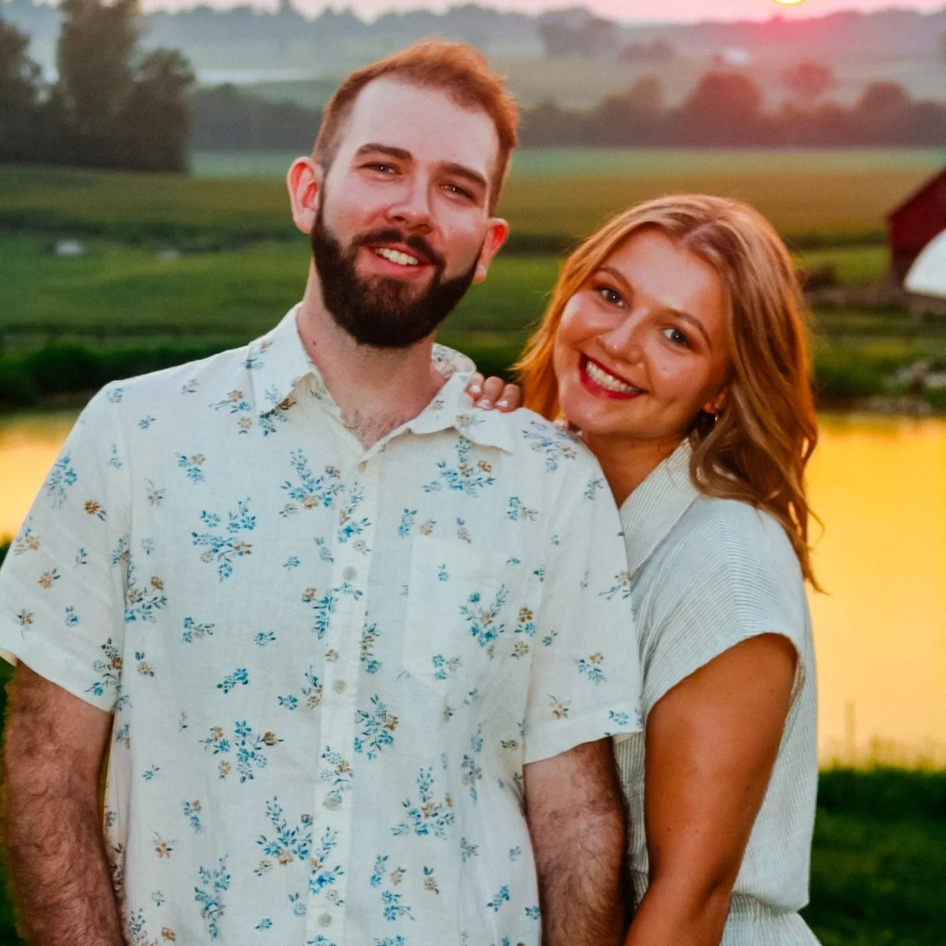 A smiling couple standing closely together outdoors during sunset with a lake and green landscape in the background.