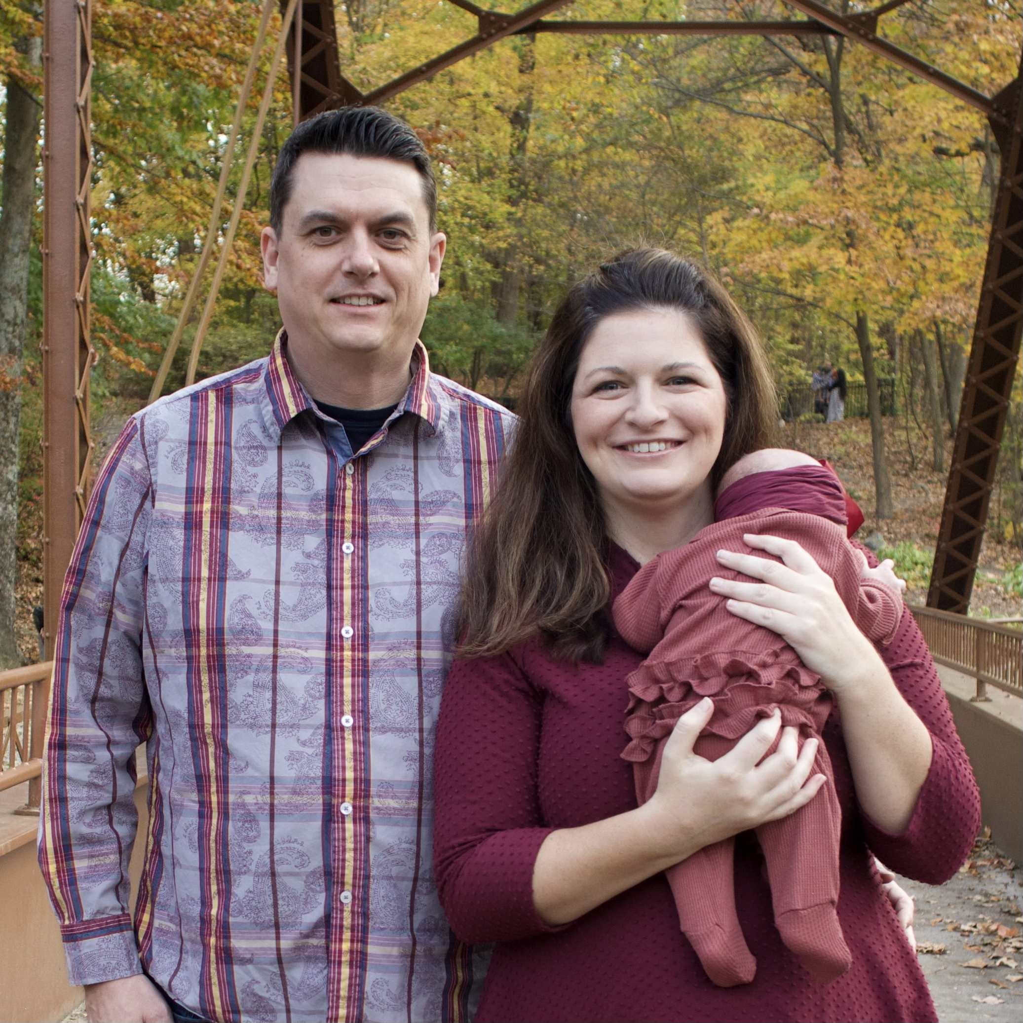 A man and a woman are standing close together outdoors in front of a bridge, with trees displaying fall foliage in the background. The man is wearing a colorful, patterned button-up shirt, and the woman is wearing a maroon top and holding a small dog.