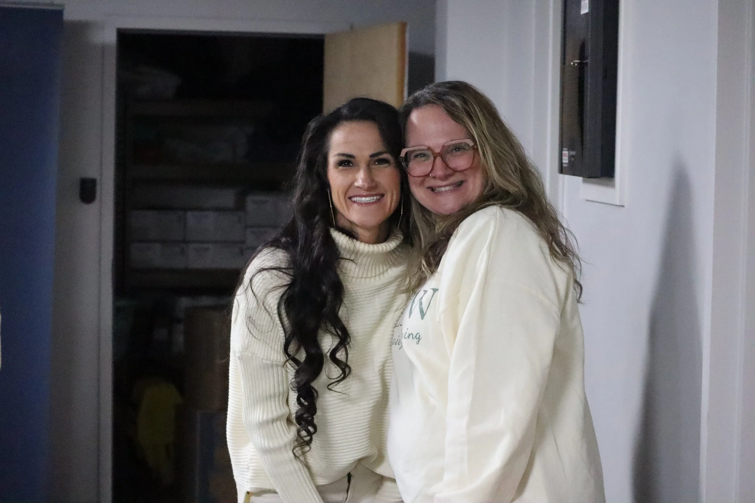 Two women standing close together indoors, smiling at the camera. One woman has long dark hair and is wearing a cream-colored sweater, the other has wavy blonde hair and glasses, wearing a light-colored top.