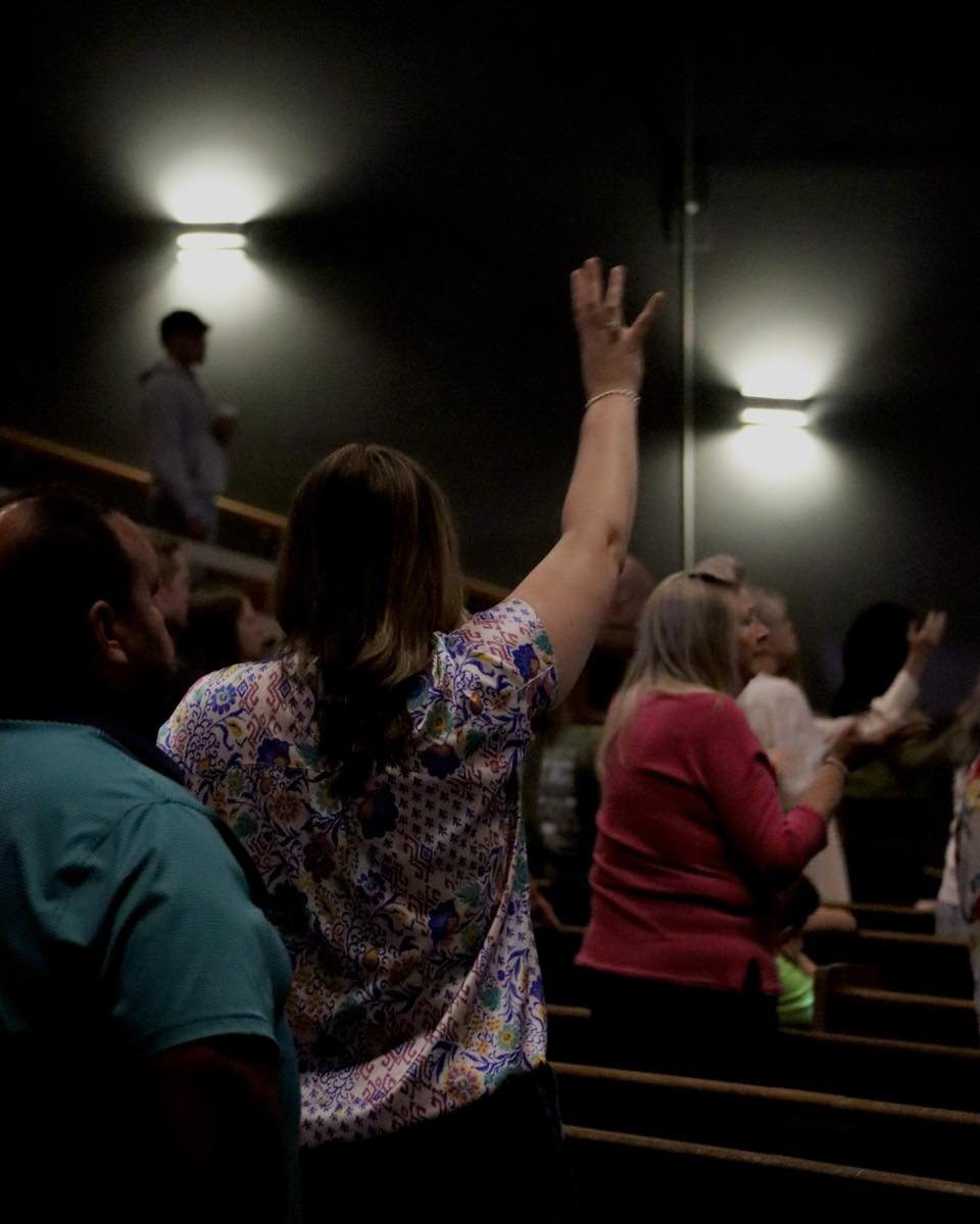 A diverse group of people in a dark auditorium or theater, with one woman in the foreground raising her hand to ask a question or make a comment, while others look on or prepare to speak.