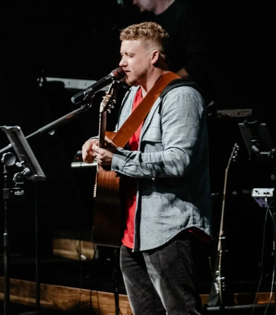 Man with curly hair playing acoustic guitar and singing into a microphone on stage with musical equipment in background.