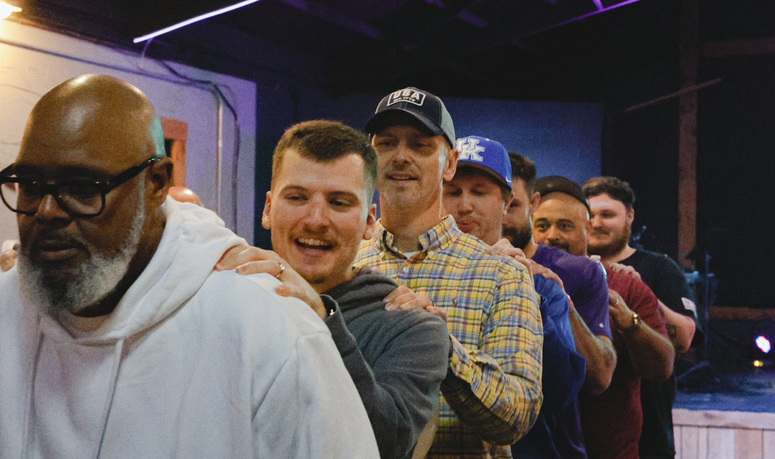 A group of men standing in a line, holding onto each other's shoulders, in a dimly lit indoor setting with neon lighting.