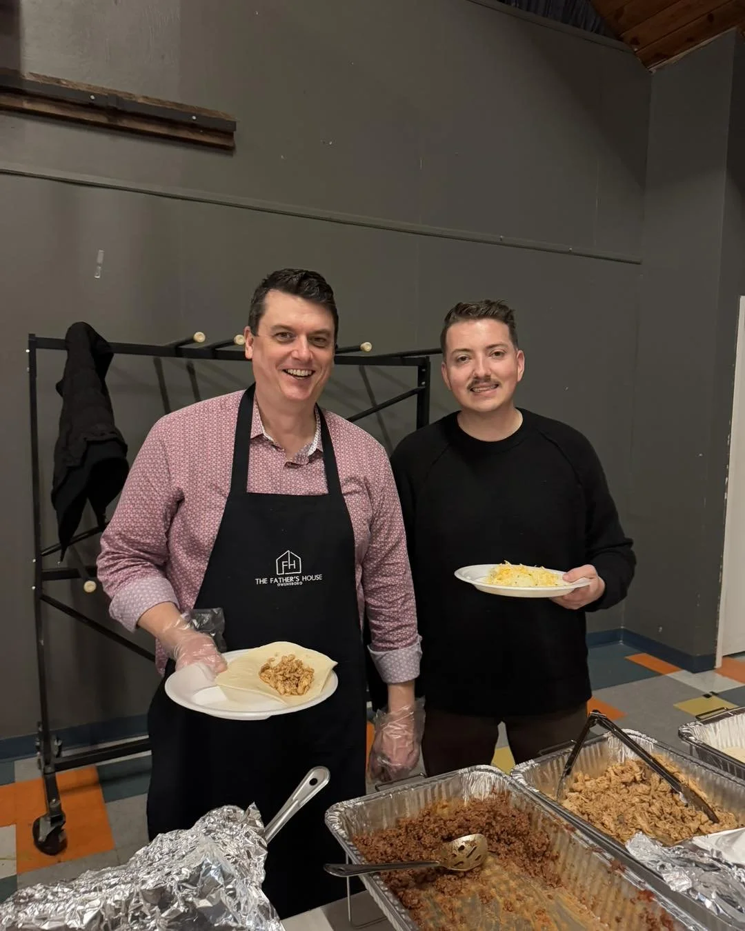 Two men standing behind a table with trays of cooked food, smiling at the camera, holding plates of food, one wearing an apron that says 'The Father's House', in a room with grey walls.