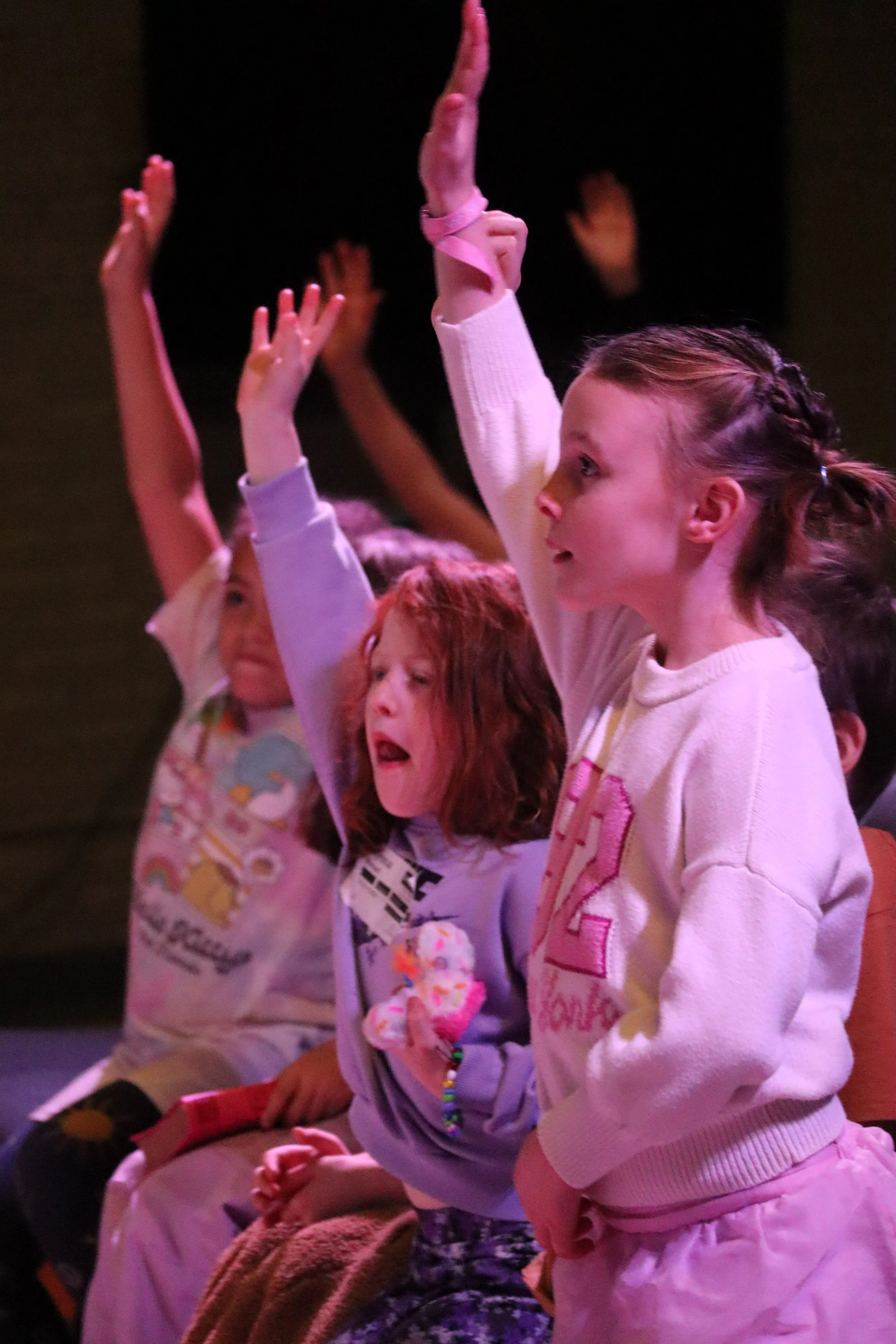 Children sitting and raising their hands, some with eyes closed or mouths open, in a dark environment with pinkish lighting.