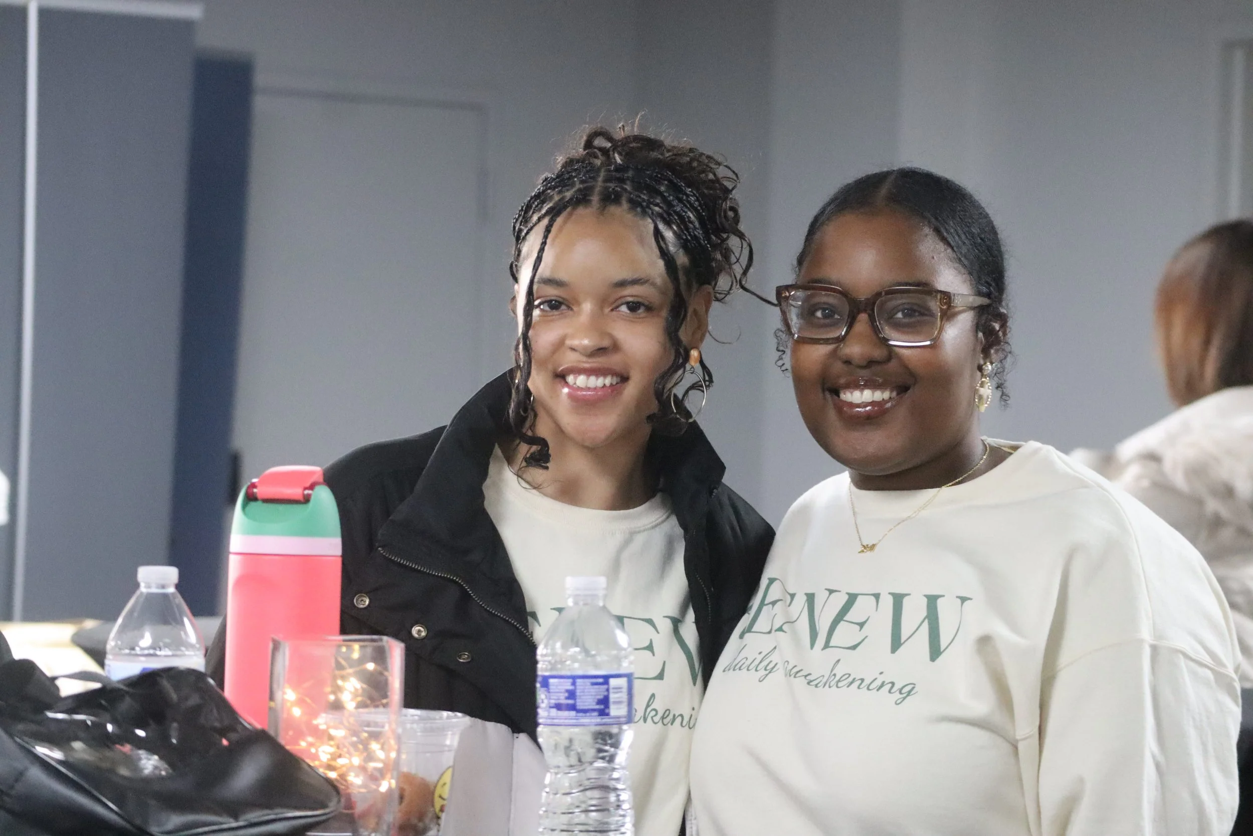 Two smiling women, one with glasses, sitting at a table with water bottles, a pink thermos, and a black bag. They are indoors, and there is a blurred background.