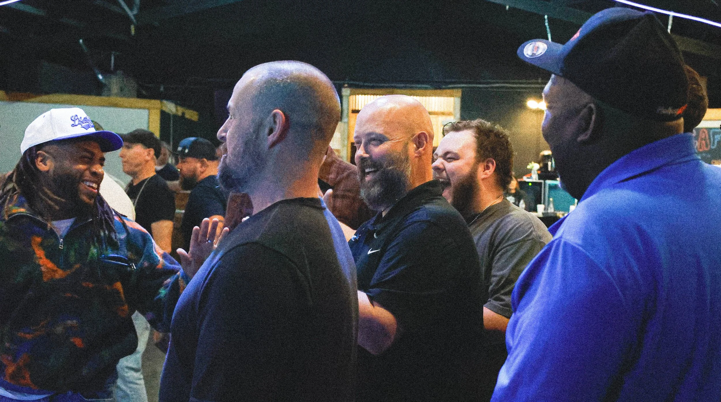 Group of people standing and talking in a dimly lit indoor setting, smiling and engaging with each other.