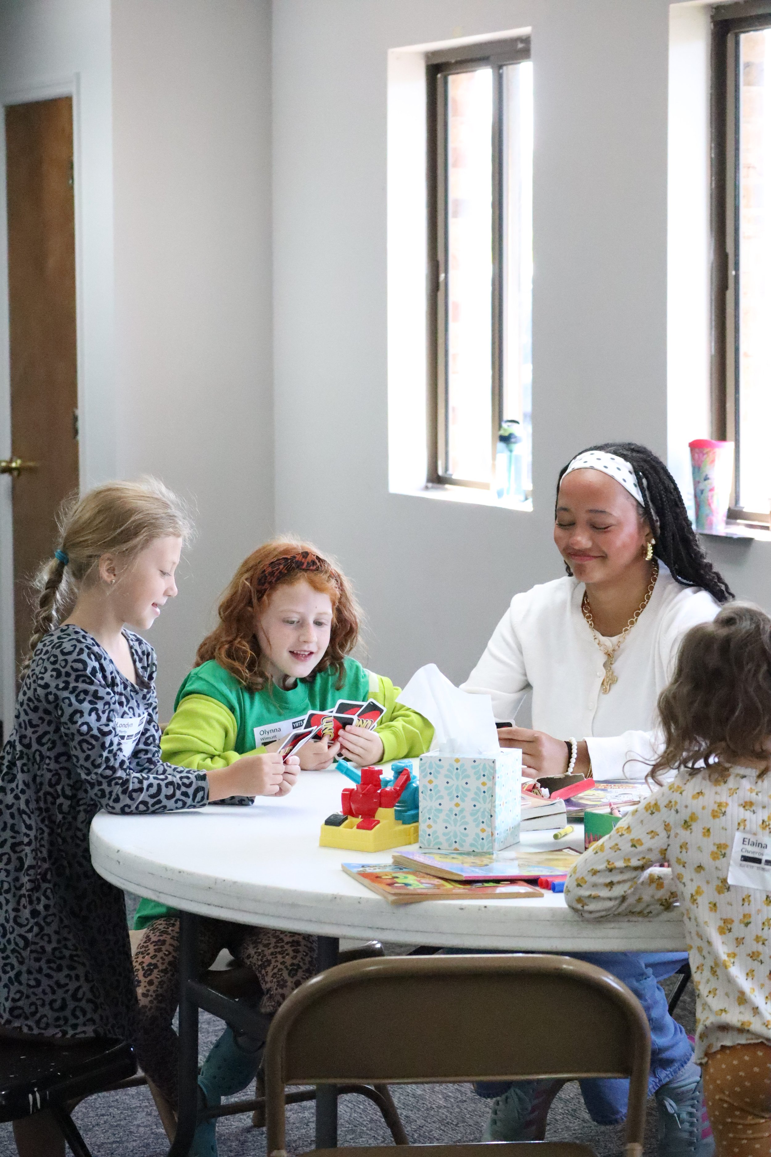 A group of children and a woman are gathered around a table inside a room, engaging in an activity. The children are holding cards, and the woman is smiling while interacting with them. There are toys, books, and a tissue box on the table.