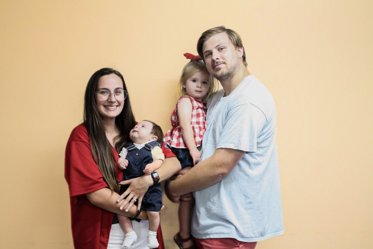 Family portrait of a woman with long dark hair and glasses, a man with short light hair, and two young girls, all standing against a light yellow wall, smiling and looking at the camera.