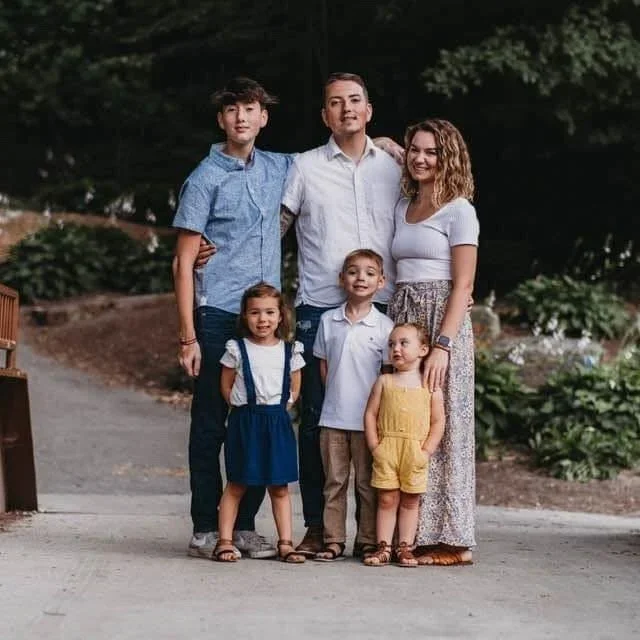 A family of six standing together outdoors, smiling for a photo, with trees and plants in the background.