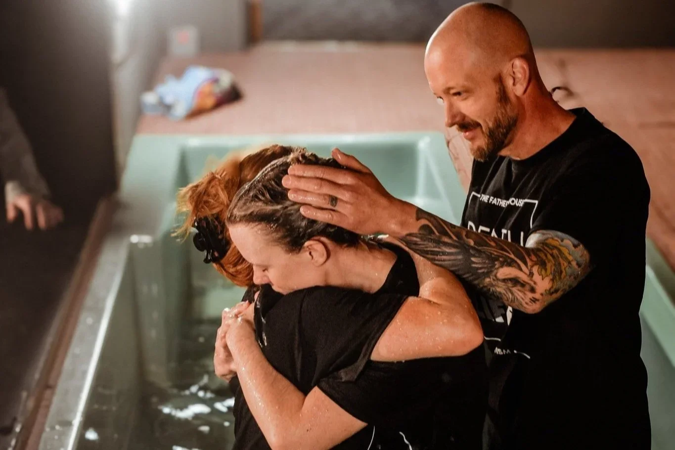 A man baptizing a woman in a church baptismal pool. The man is gently pouring water over the woman's head while she is bowed and crying. The woman has reddish hair tied back, and the man has tattoos on his arm and is wearing a black shirt.