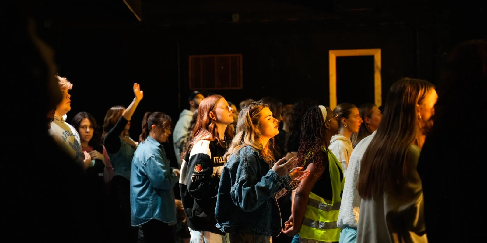 Group of young adults in a dark room, some with eyes closed and faces uplifted, participating in a concert or worship event, illuminated by warm lighting.