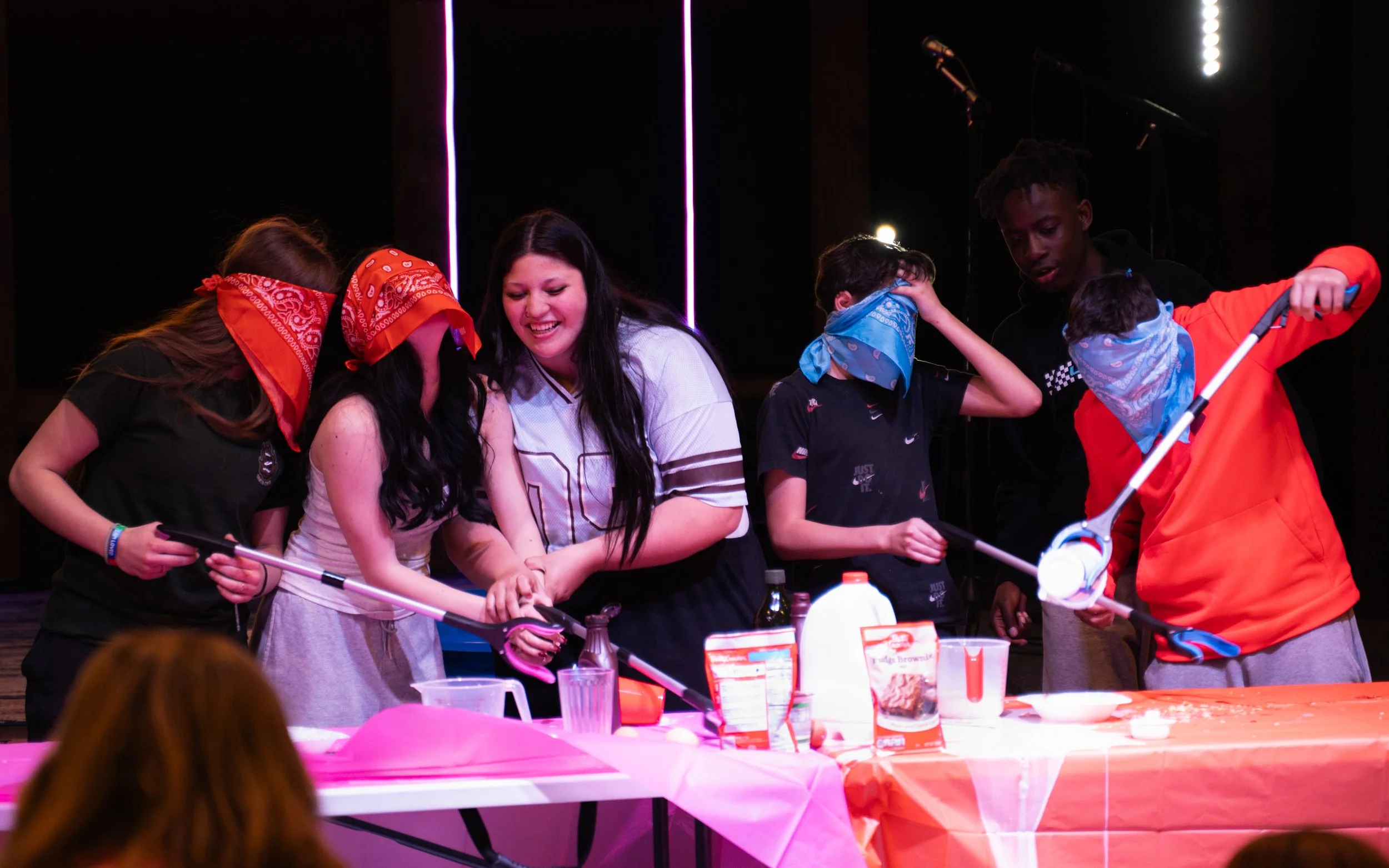Group of young people wearing bandanas, gathered around a table with baking ingredients, engaged in a baking activity in a dimly lit room with purple neon lights.