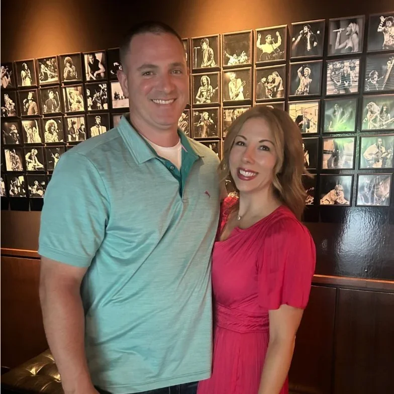 A smiling man and woman posing together at a photo gallery with black and white photos of musicians and performers on the wall behind them.