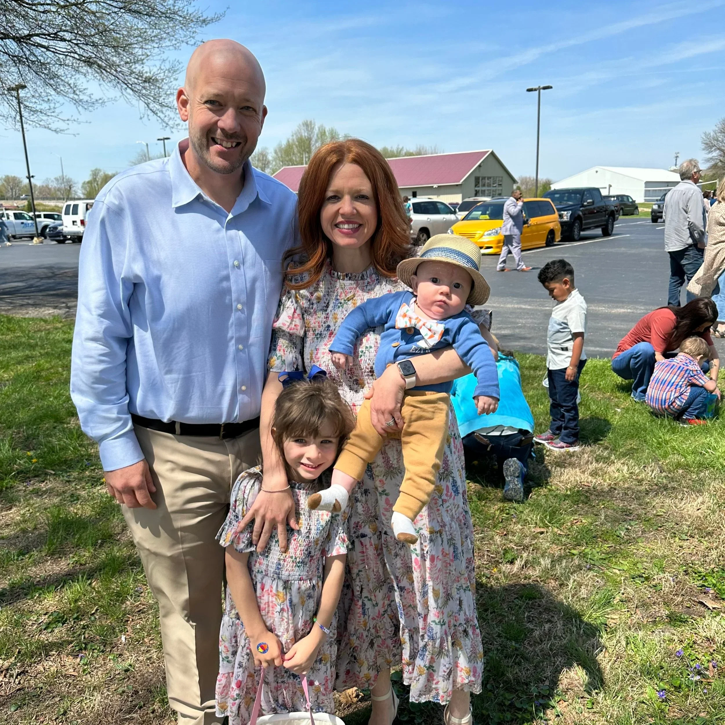 A man with a bald head and a woman with red hair standing outdoors, holding a young child in front of a parking lot with several cars and small buildings. The sky is blue with some clouds, and there are trees and people in the background.
