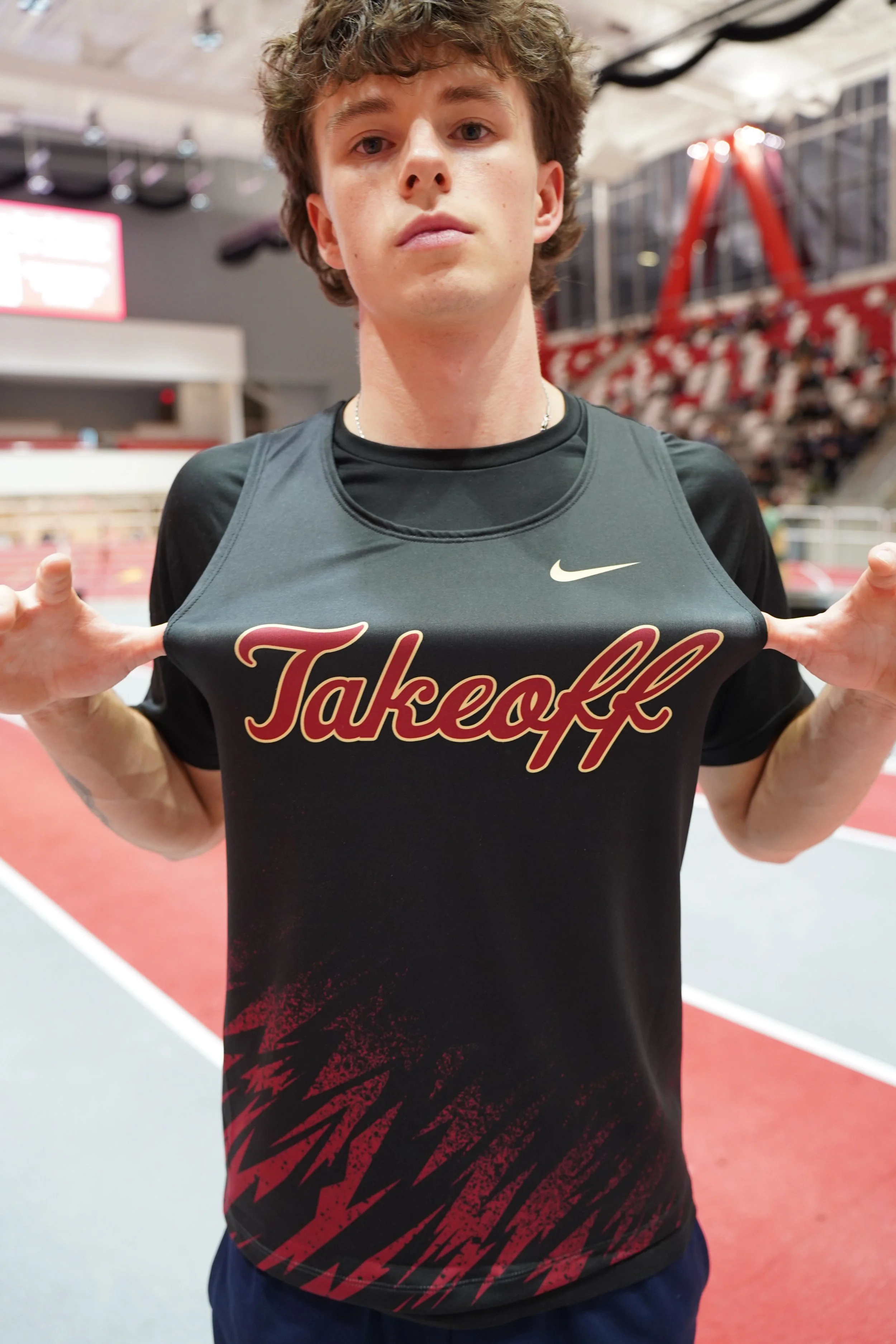 Young man holding up his sports shirt with both hands, displaying the word 'Takeoff' on the front inside an indoor sports facility with a track and blurred background.