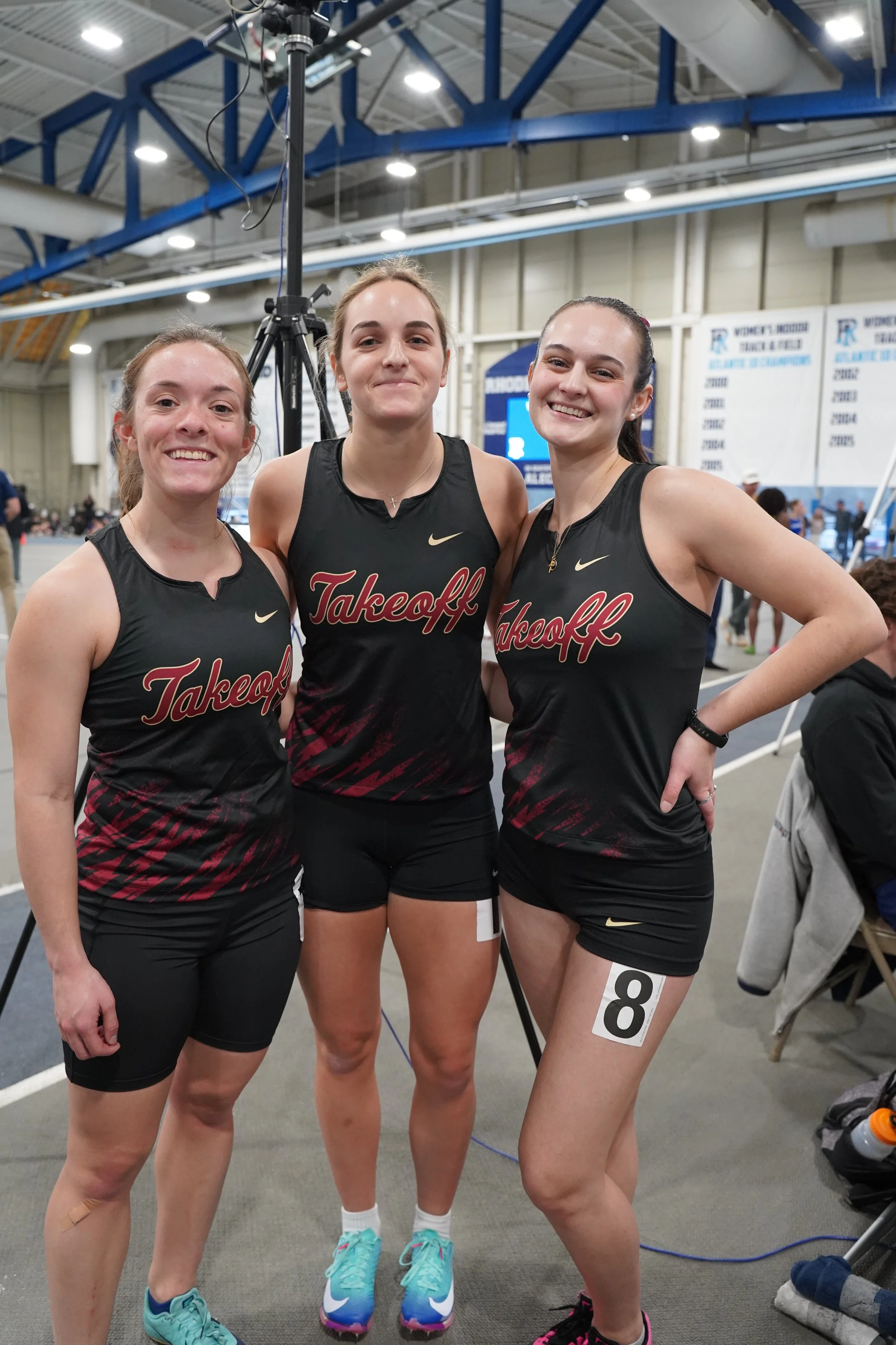 Three young female athletes in black sports uniforms with 'Takeoff' branding posing together indoors at a track and field event, smiling after a race.