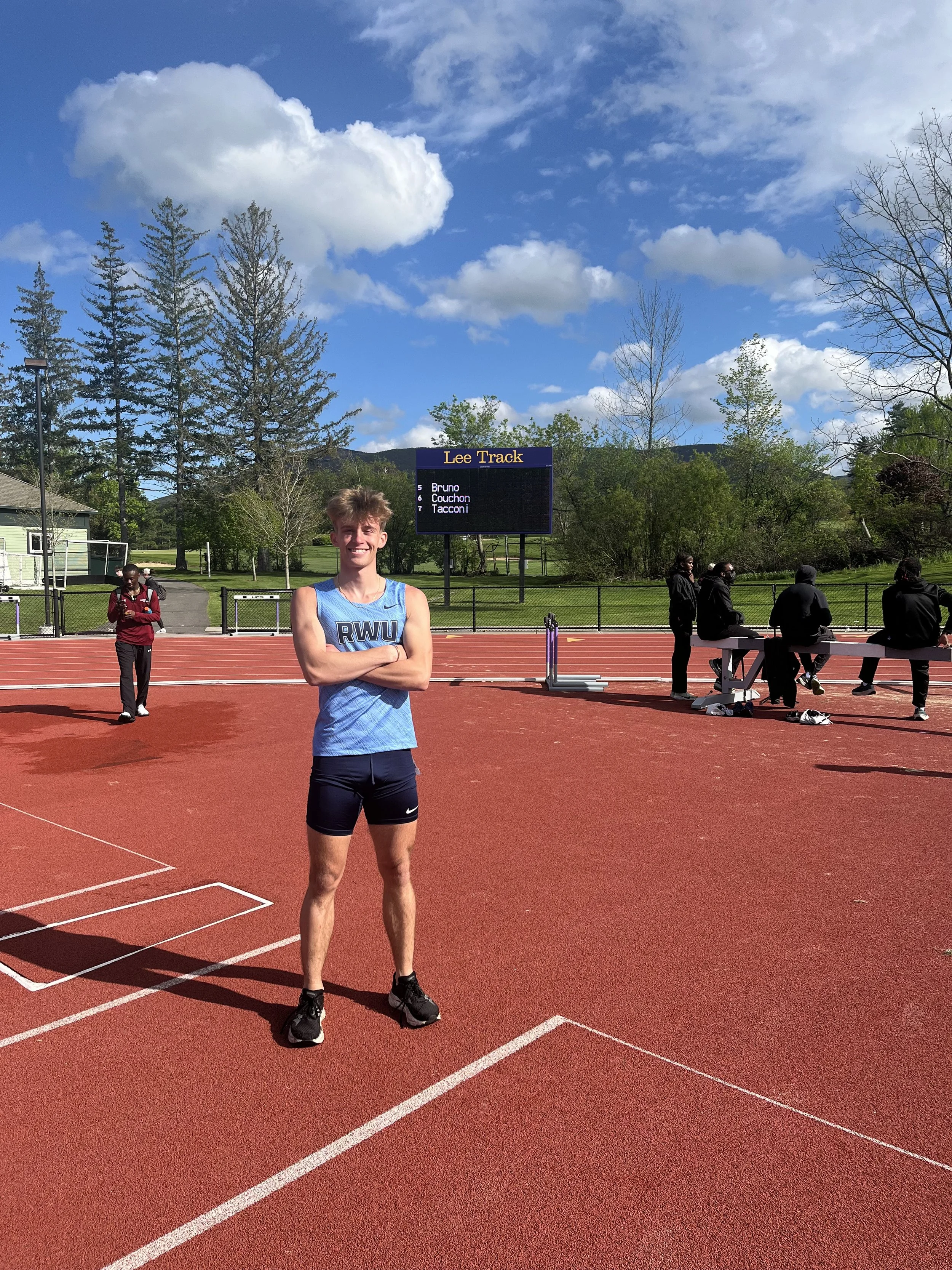 Young male athlete standing on a red track field with his arms crossed, smiling at the camera. In the background, there are trees, a fence, a scoreboard displaying names of athletes, and a partly cloudy sky.
