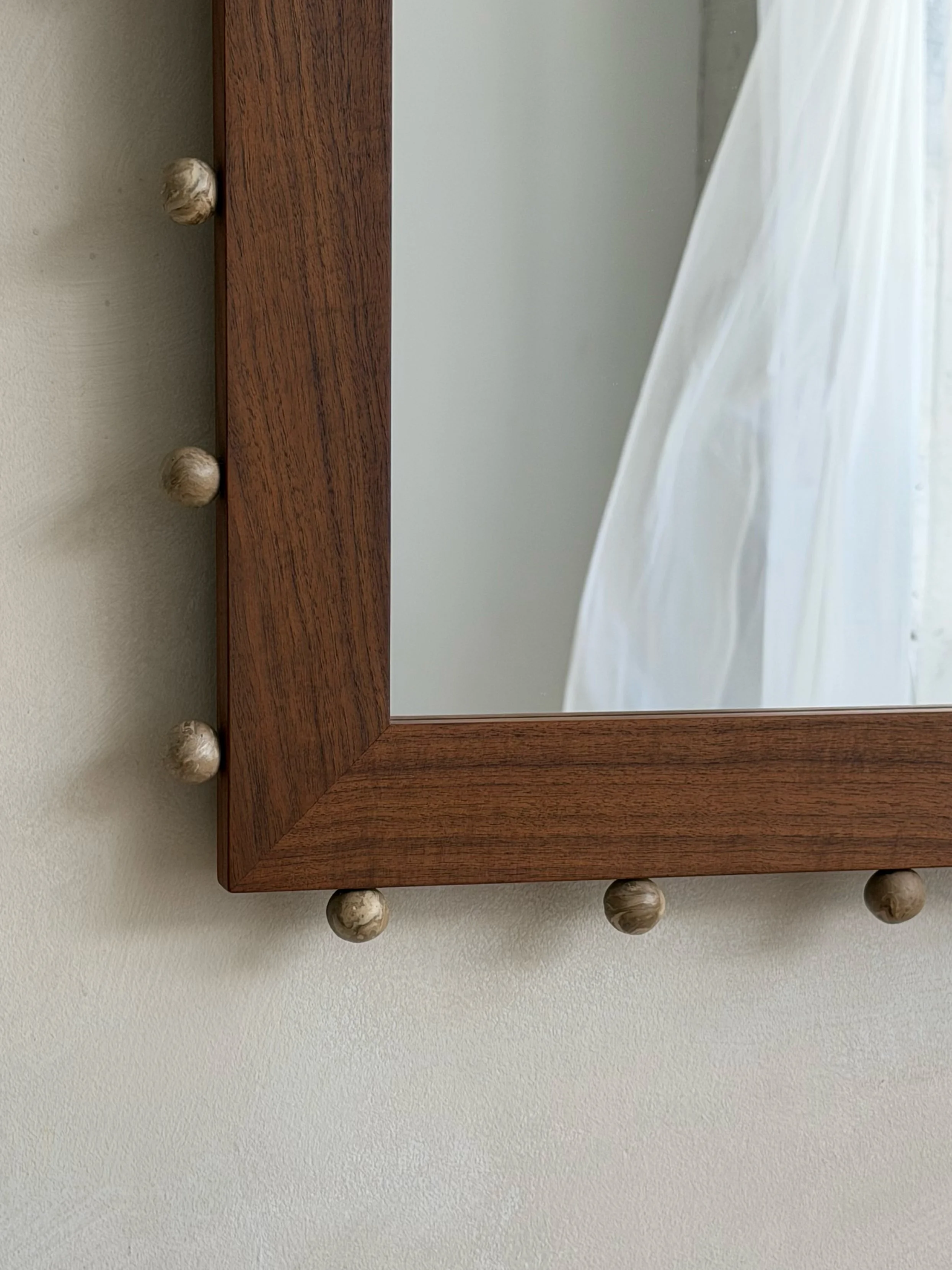 Wooden-framed mirror with round ceramic ball embellshiments on the border, hanging on a beige wall, reflected curtain and wall.