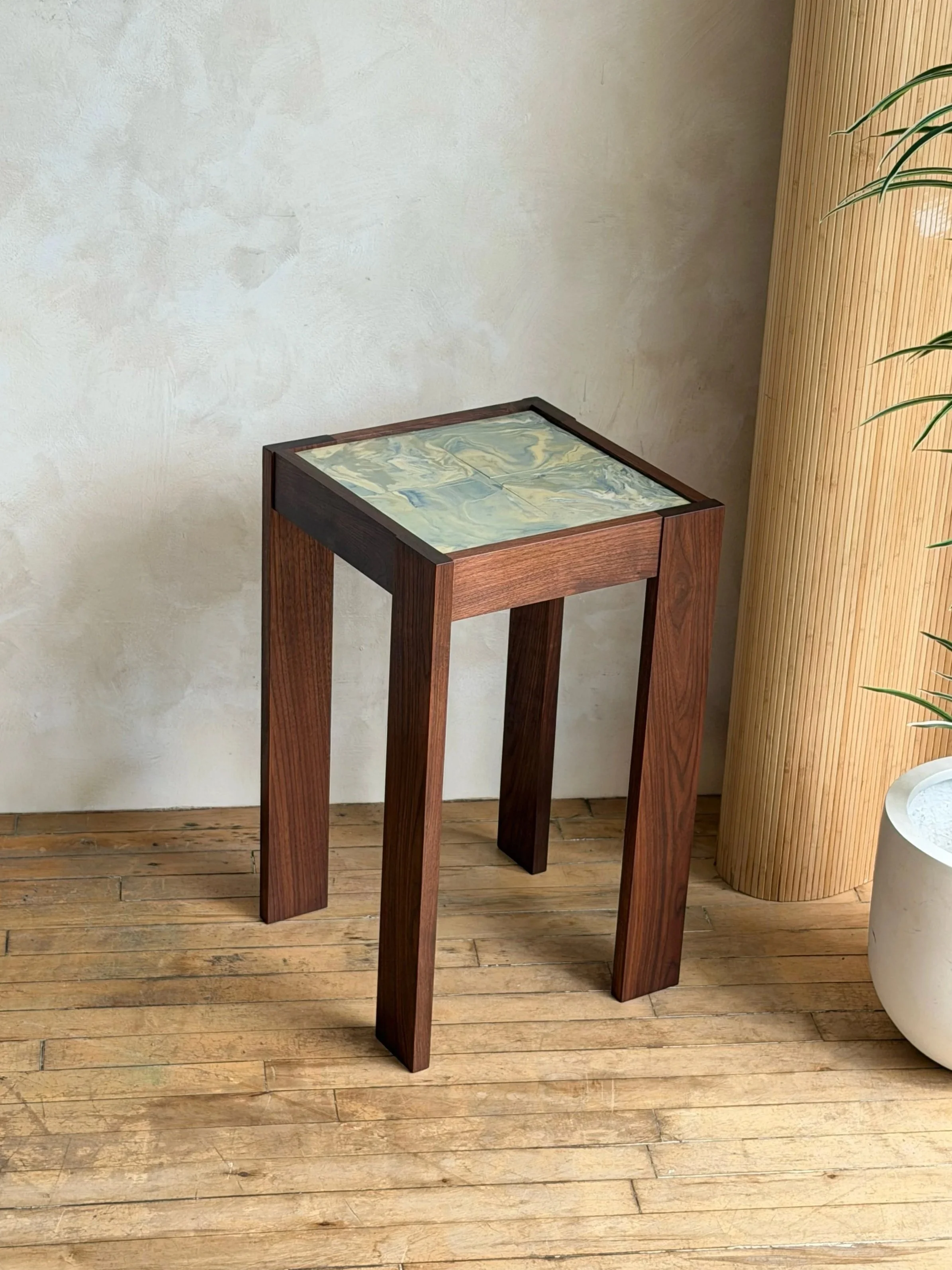 A wooden table with a marbled ceramic top, standing on four legs in a room with wooden flooring and a textured wall.