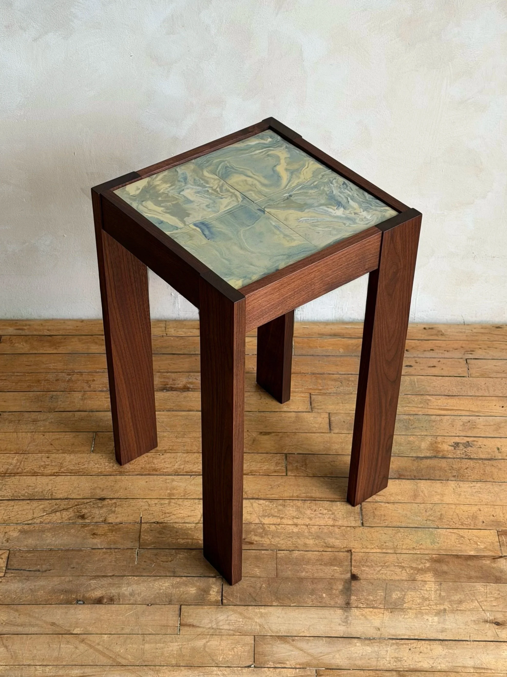 A small wooden table with a square marbled ceramic top featuring a swirling pattern of greens and tans. The table is on a wooden floor against a plain white wall.