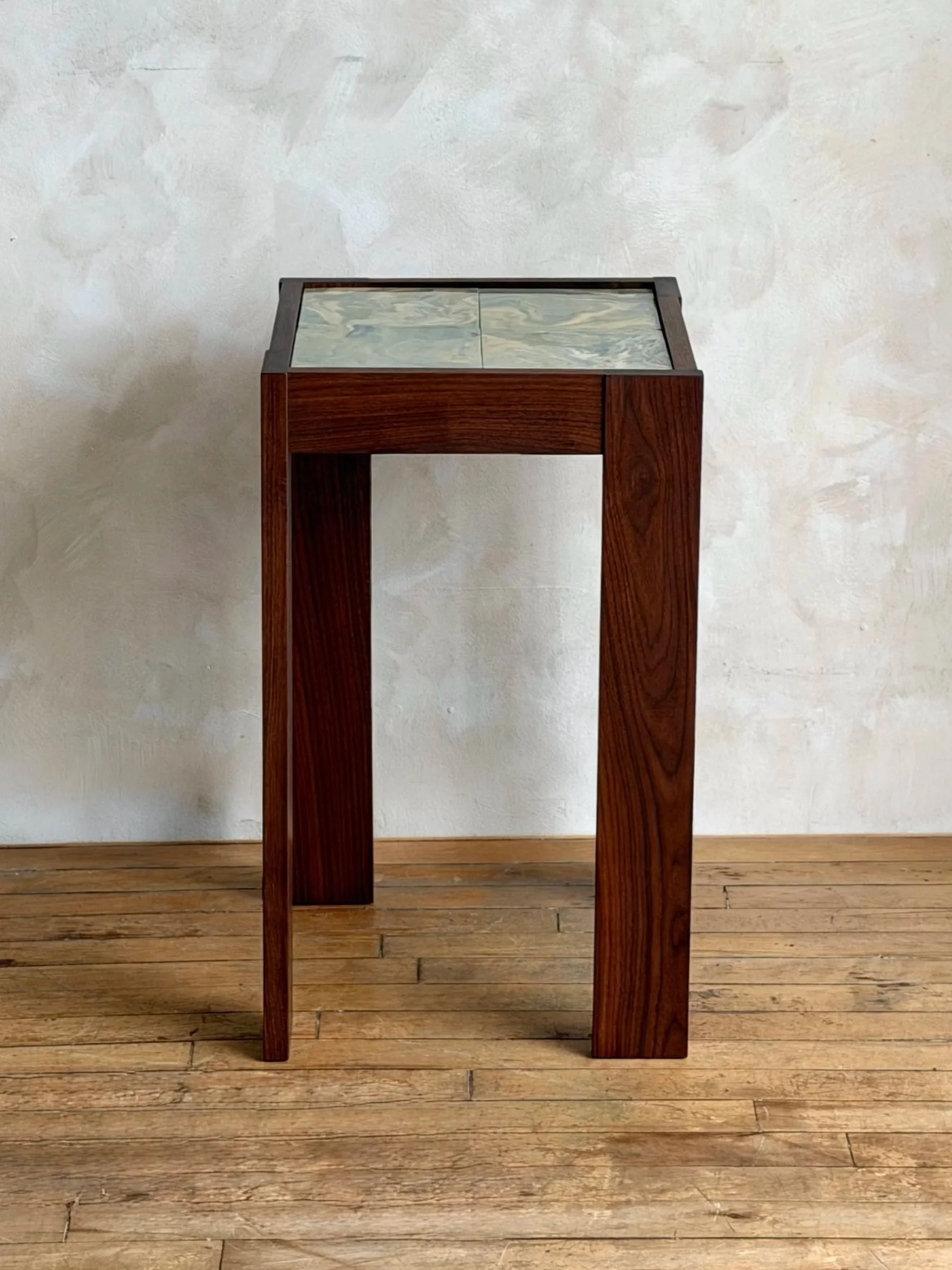 A wooden table with a marbled ceramic top, placed against a white textured wall and on a wooden floor.