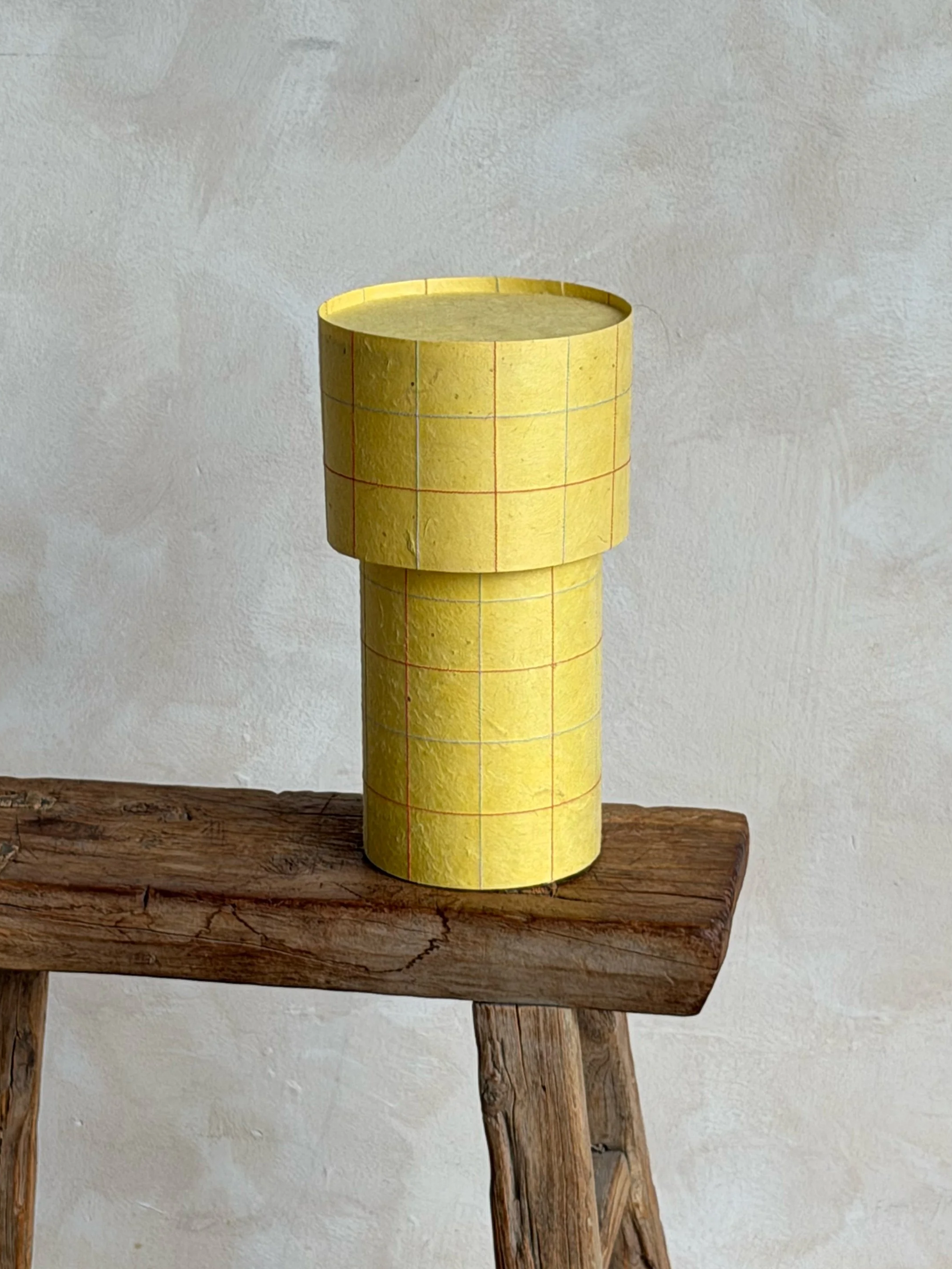 A yellow cylindrical lamp with red grid lines on top of a weathered wooden surface, against a white textured wall.
