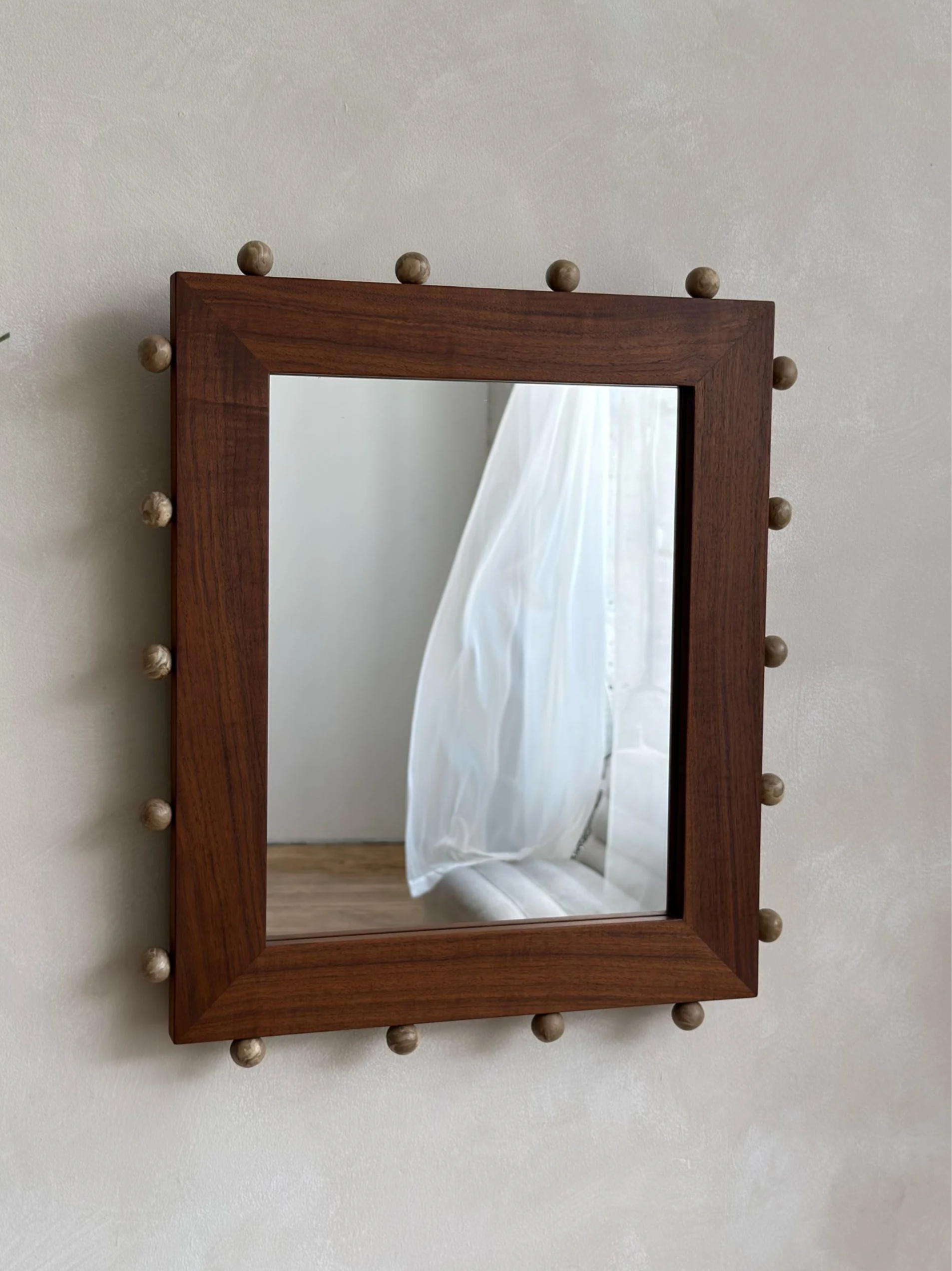 Wooden-framed mirror with ceramic ball-shaped embellishments on the edges, reflecting a white curtain and part of the wall.