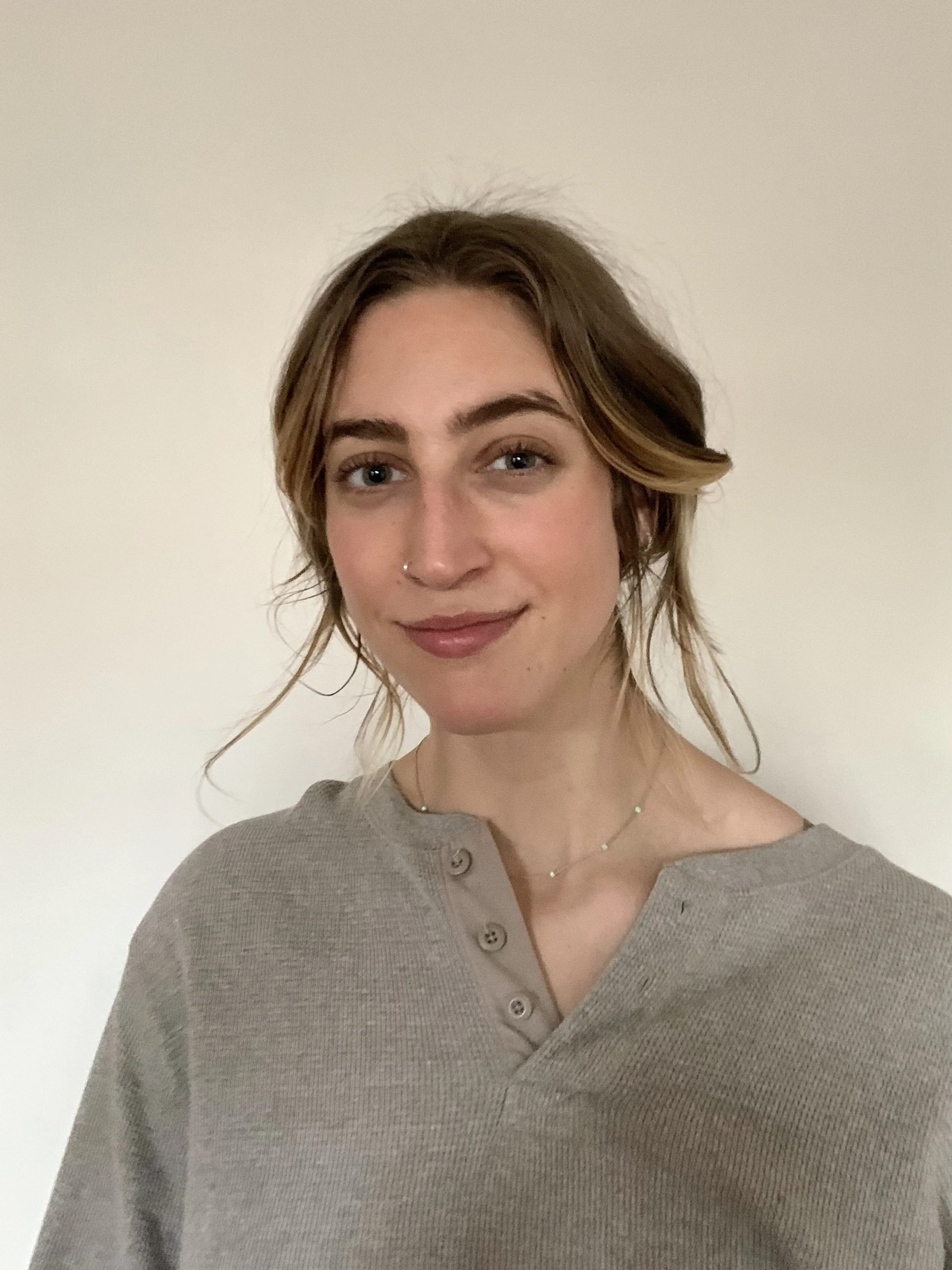 Headshot of a young woman with shoulder-length wavy brown hair and a nose ring, wearing a beige button-up sweater with a v-neck, standing against a plain off-white wall.