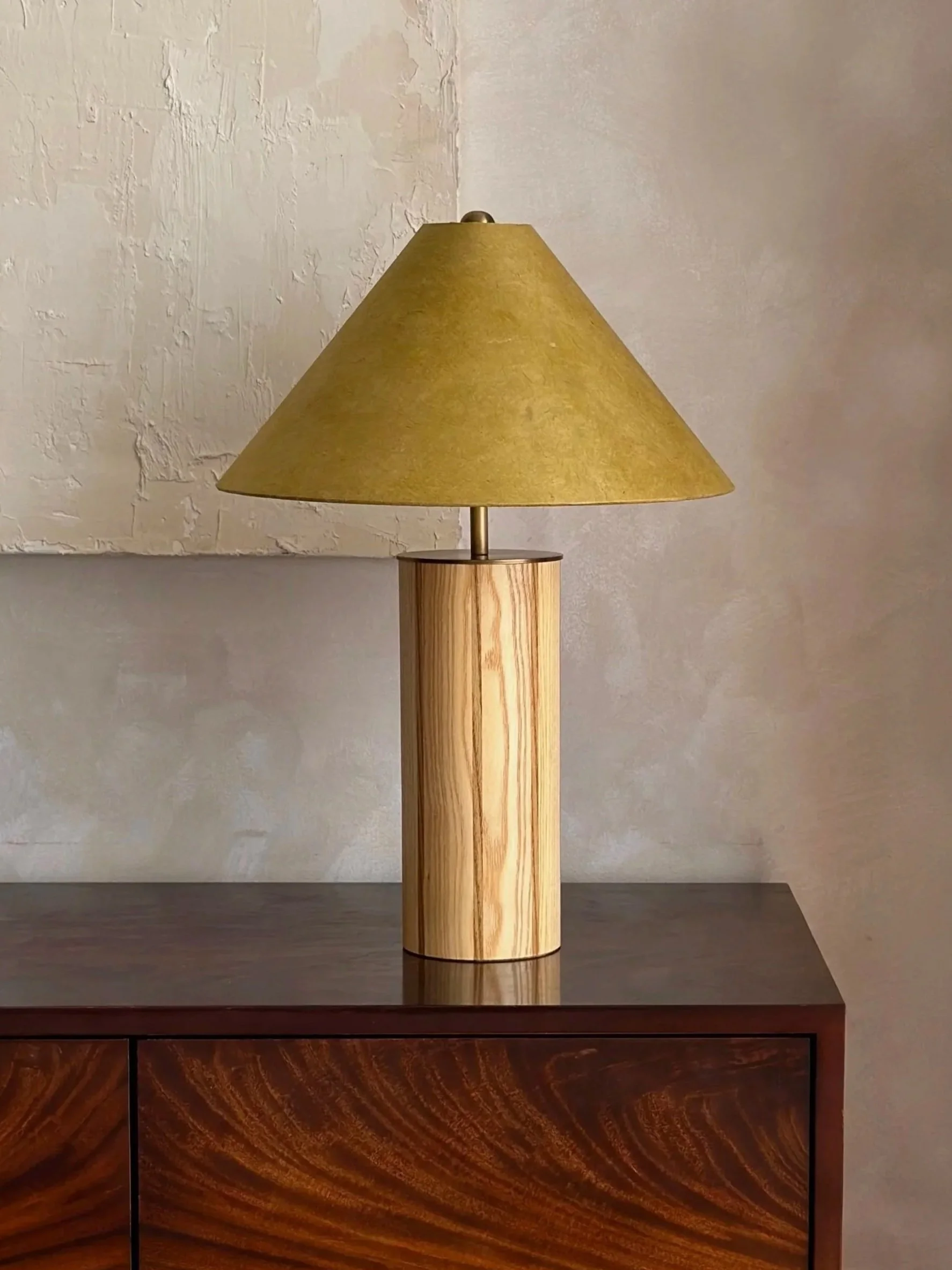 A table lamp with a green-colored conical lampshade and a cylindrical wooden base, placed on a dark wooden table against a textured wall background.